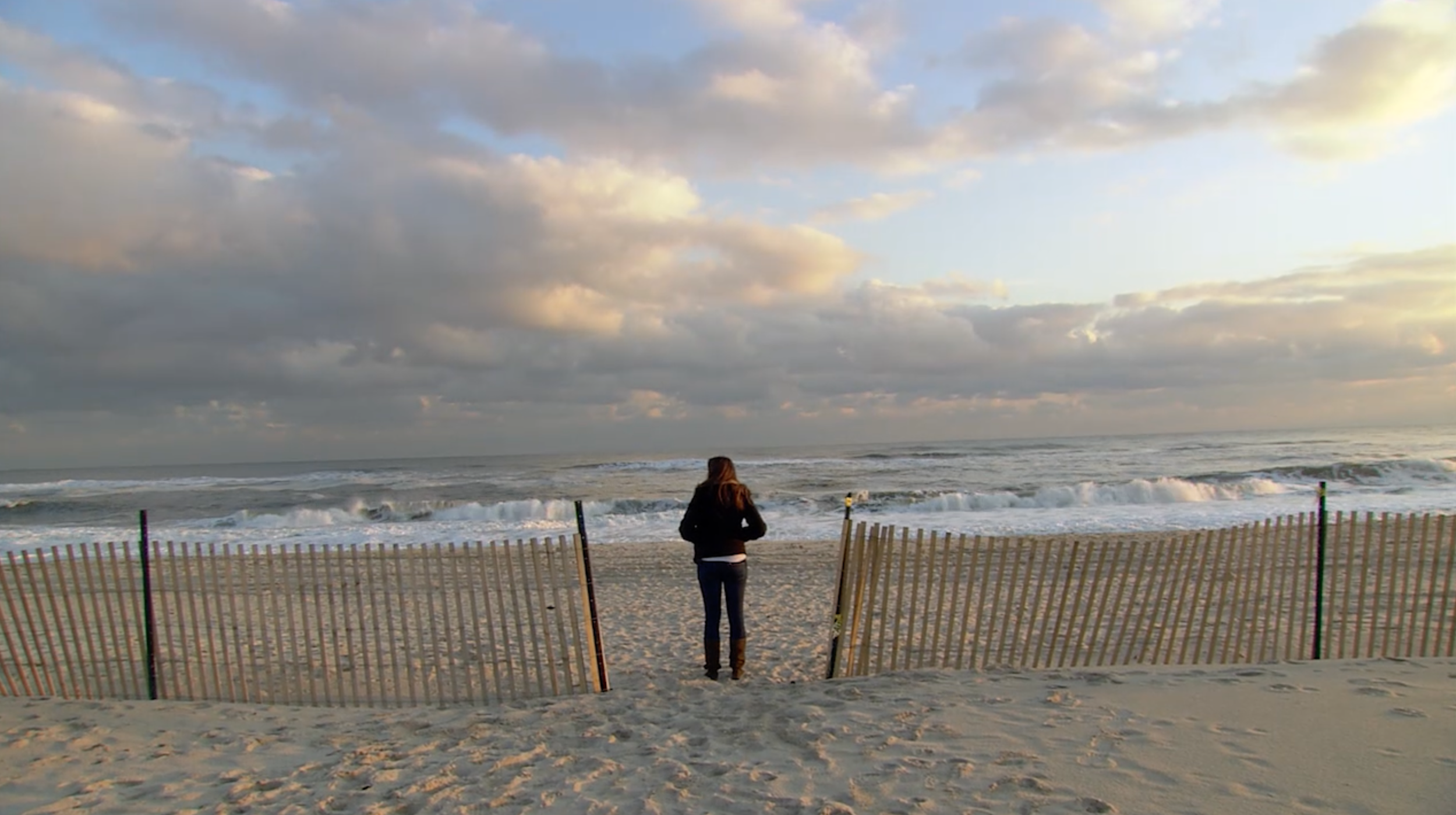 A person standing on a sandy beach looking at the ocean with waves and cloudy sky in the background. A fence is visible in the foreground.