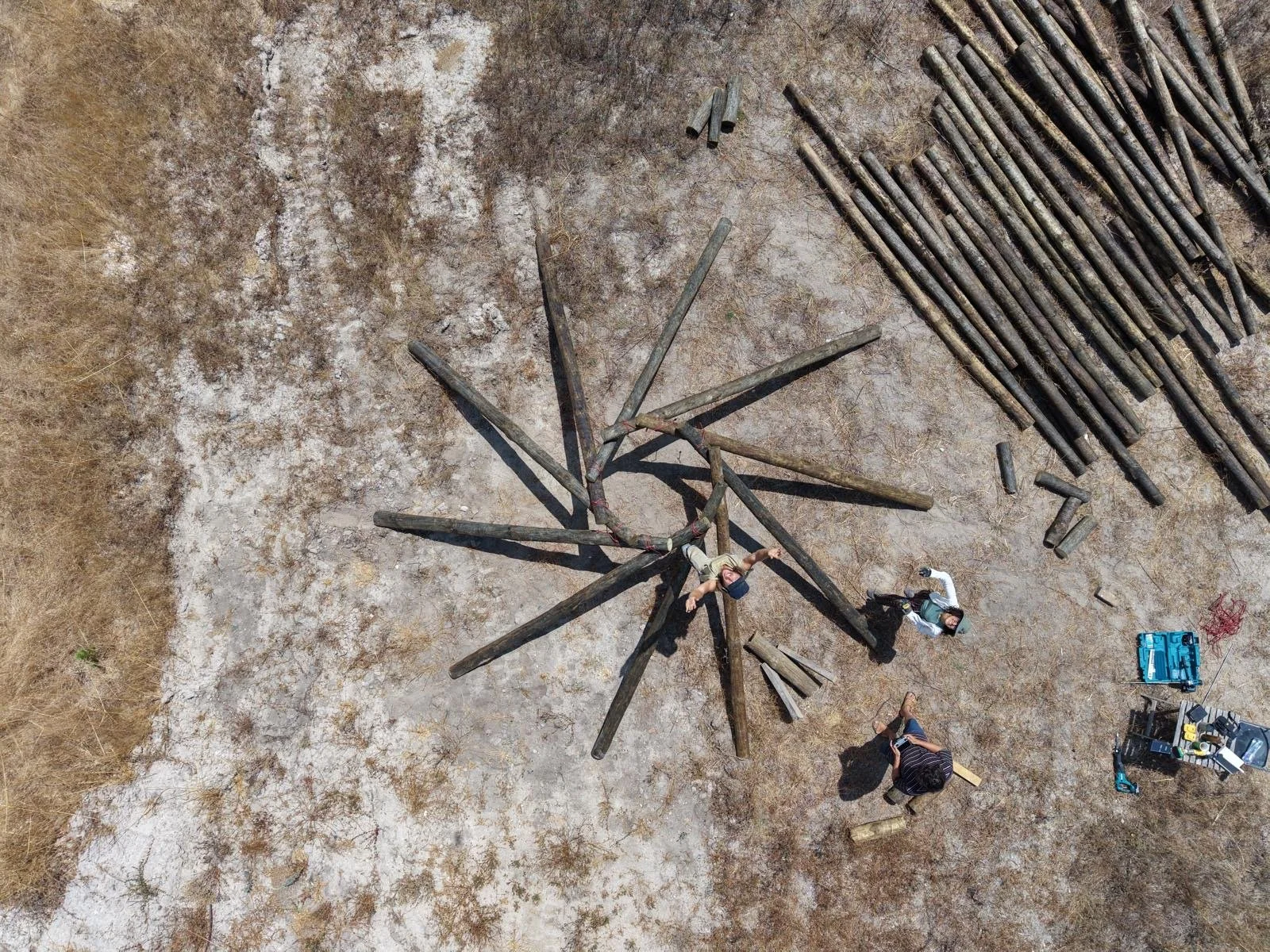 Aerial view of men building a large wooden structure, a reciprocal roof structure outdoors on a dusty ground, with tools and finished logs nearby.