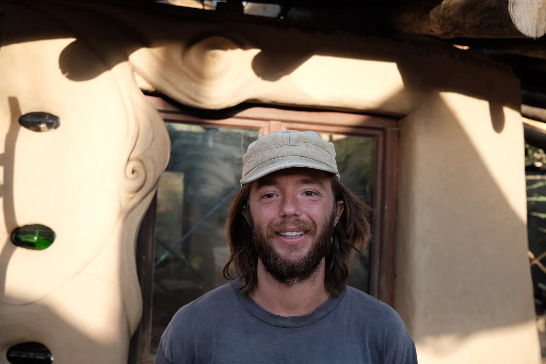 A man with long hair and a beard wearing a beige cap and a gray shirt, smiling at the camera, standing in front of a rustic structure with curved wooden beams and decorative elements.