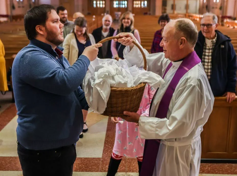A priest in a white robe with a purple stole receives a basket from a man in a blue sweater during a church ceremony, with people gathered in the background.