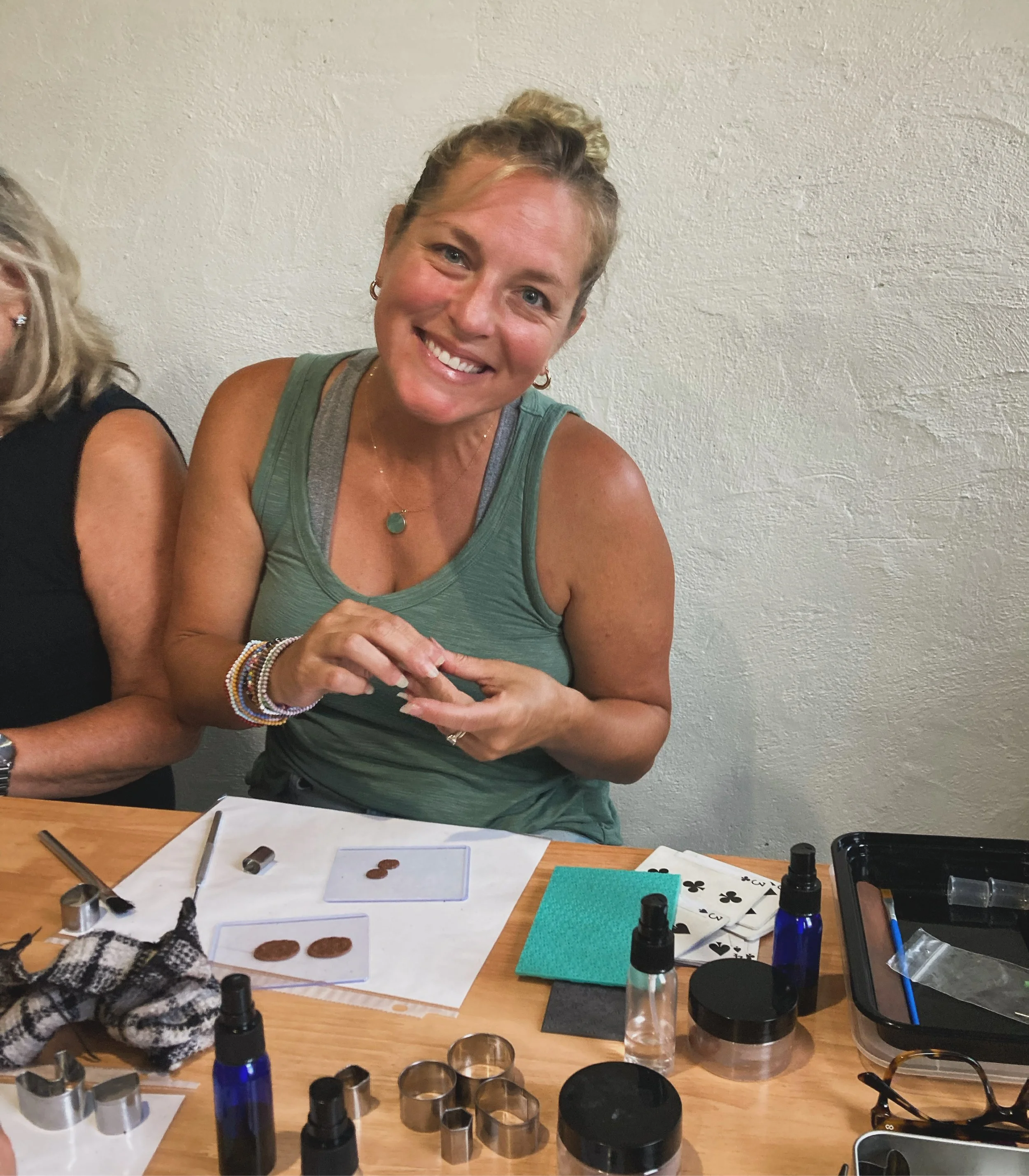 A woman with blonde hair tied back smiling and sitting at a table with various jewelry-making tools and materials, including small bottles, metal rings, and paper with coin designs.