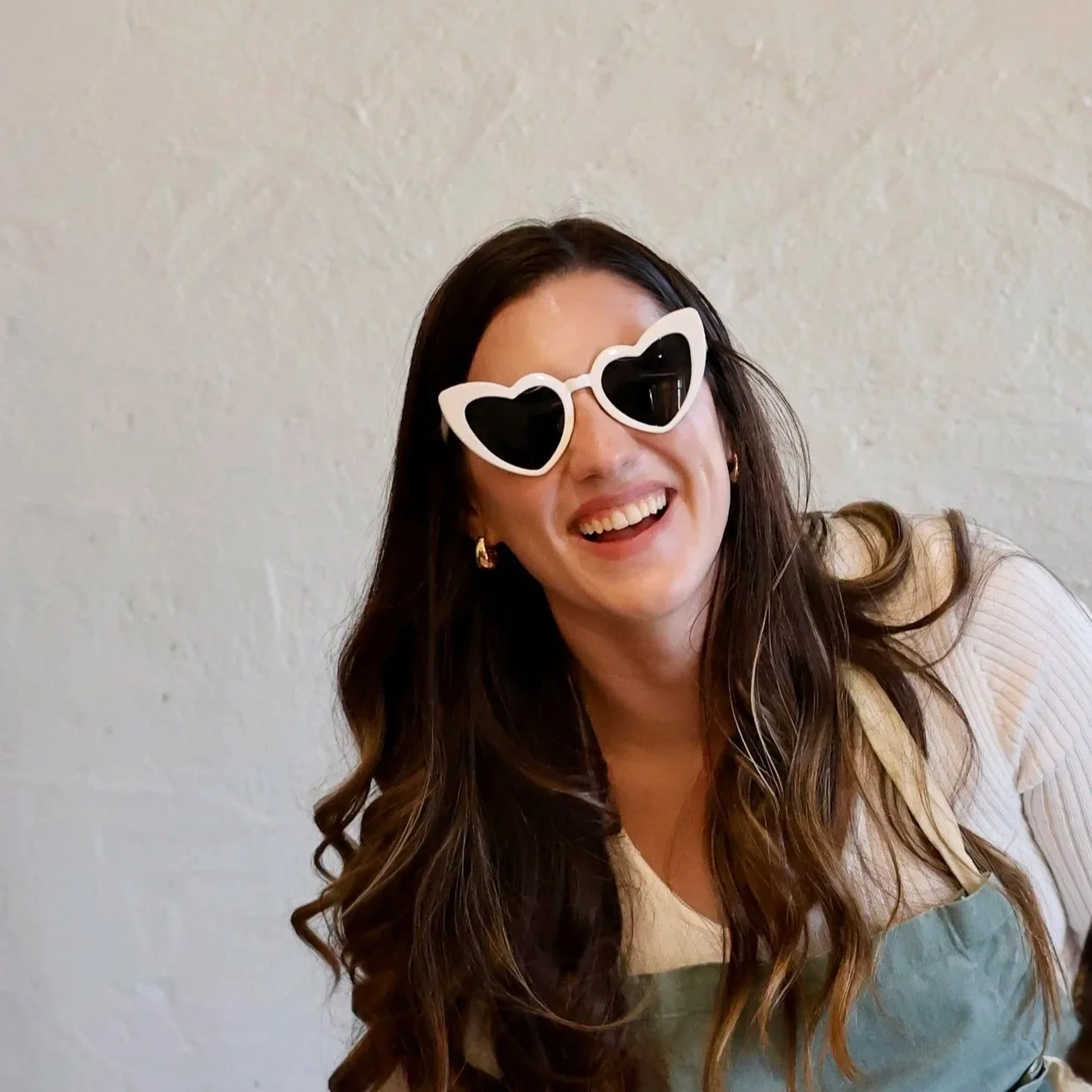 A woman with long dark hair smiling with heart-shaped sunglasses and gold hoop earrings, wearing a cream-colored top and green apron, against a light-colored wall.