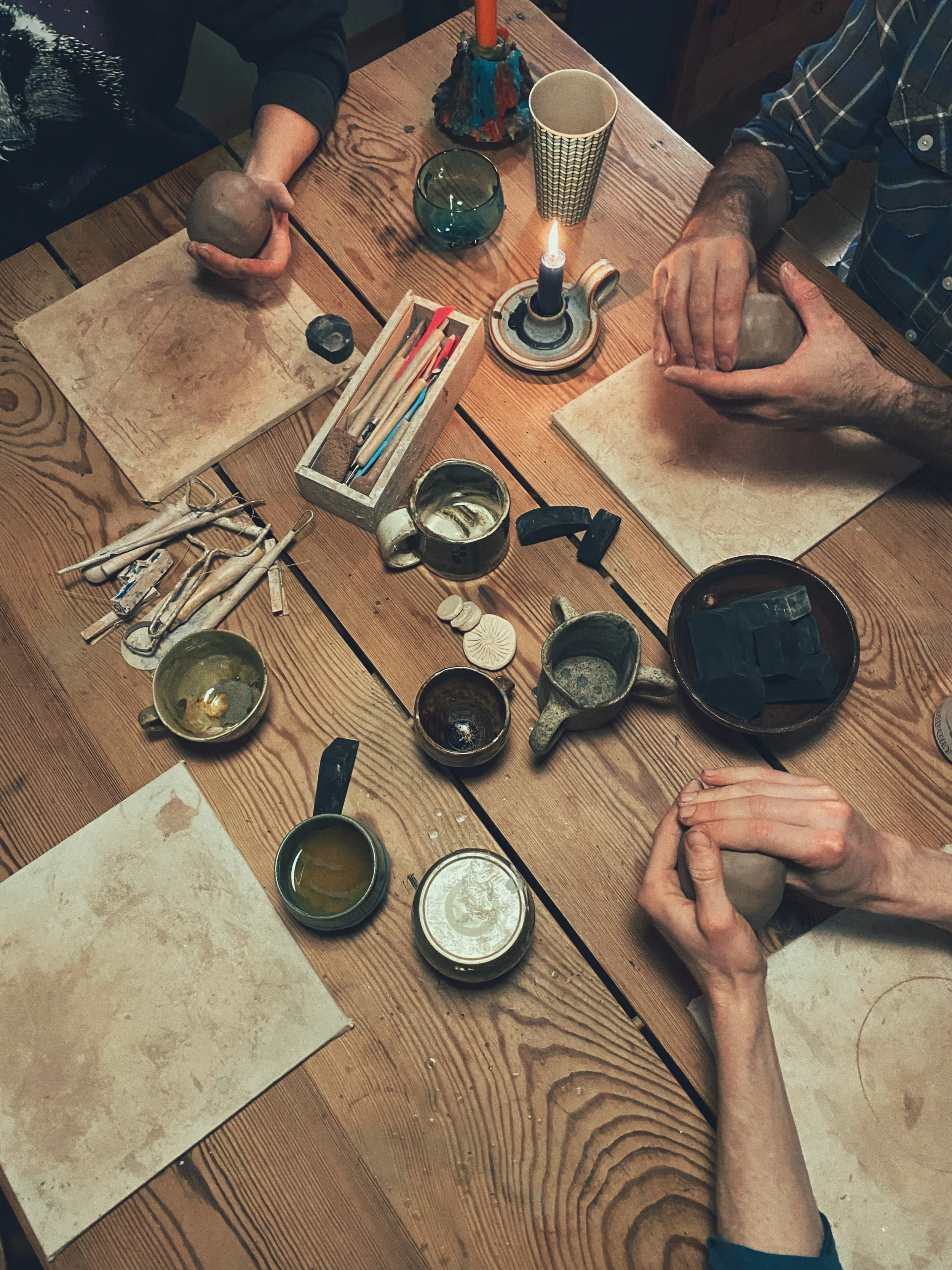 People working on pottery at a wooden table with tools, bowls of clay, a candle, and cups.