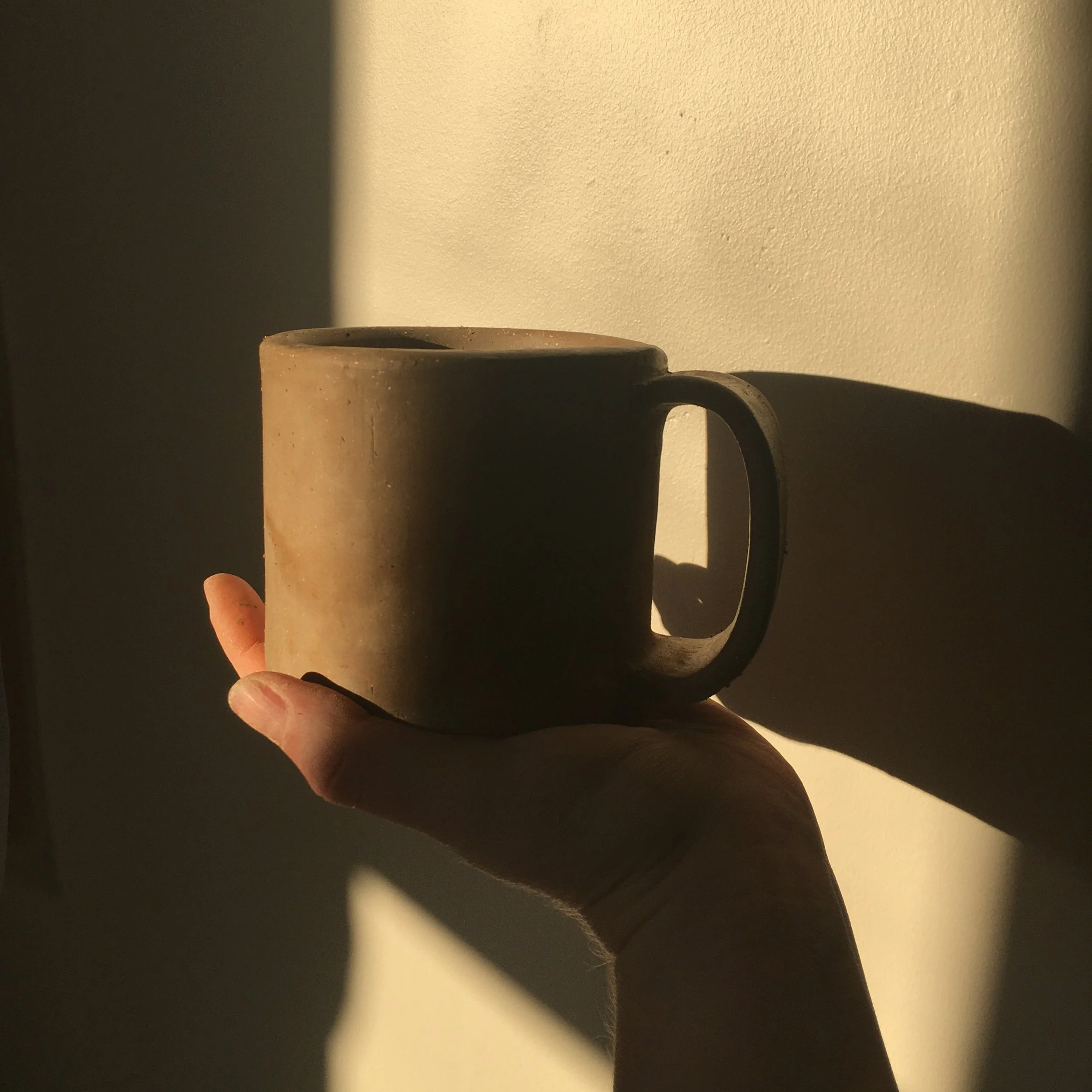 Close-up of a hand holding a beige ceramic mug with a handle, against a backdrop of a plain, textured wall with sunlight creating shadows.