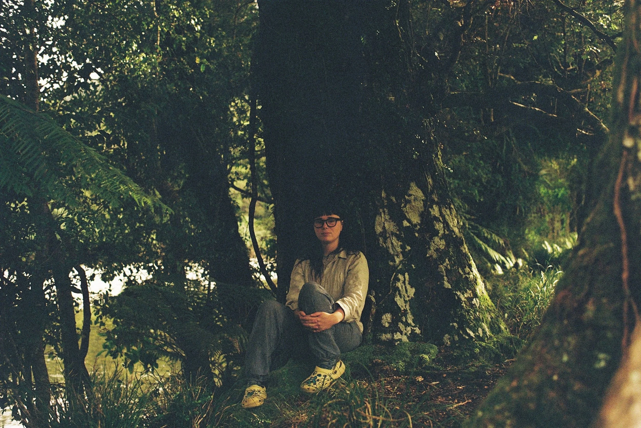 A woman with glasses and dark hair sitting on a mossy log in a dense forest, leaning against a large tree trunk with thick branches and green leaves.