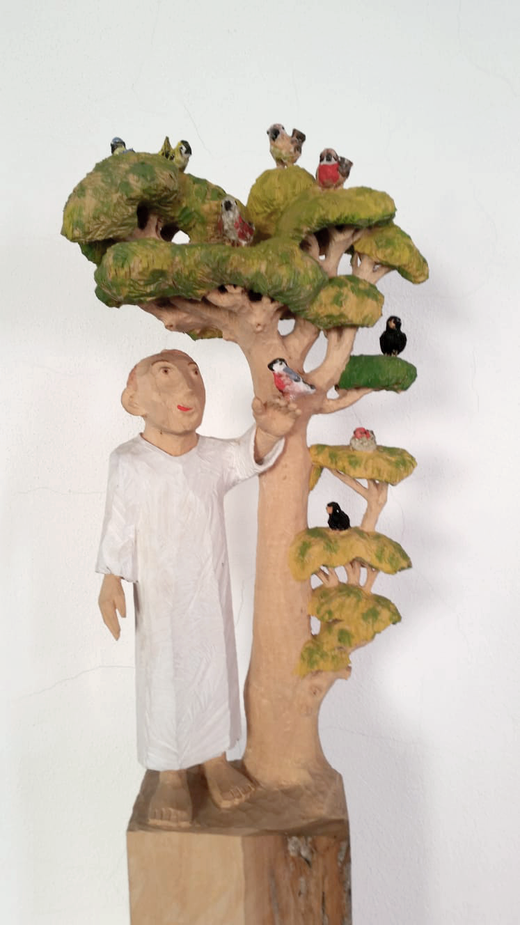 Holzfigur von Person in weißem Gewand neben einem Baum mit Vögeln