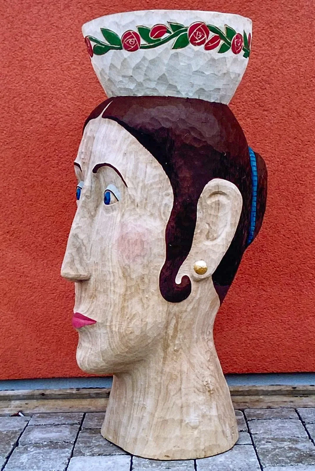 Holzskulptur eines Kopfes mit einer bemalten Schüssel auf dem Kopf, vor einer orangefarbenen Wand.