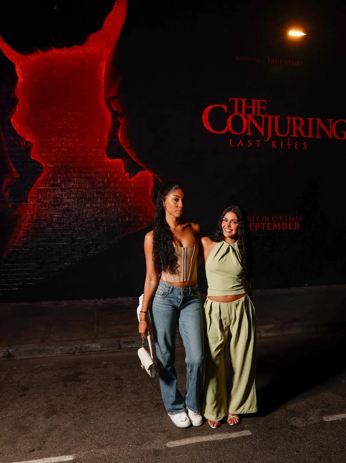 A woman with long wavy hair wearing a black sequined dress and a large silver necklace, standing in front of a black backdrop with the word "SMILE" and the number "2" repeated in white and red.