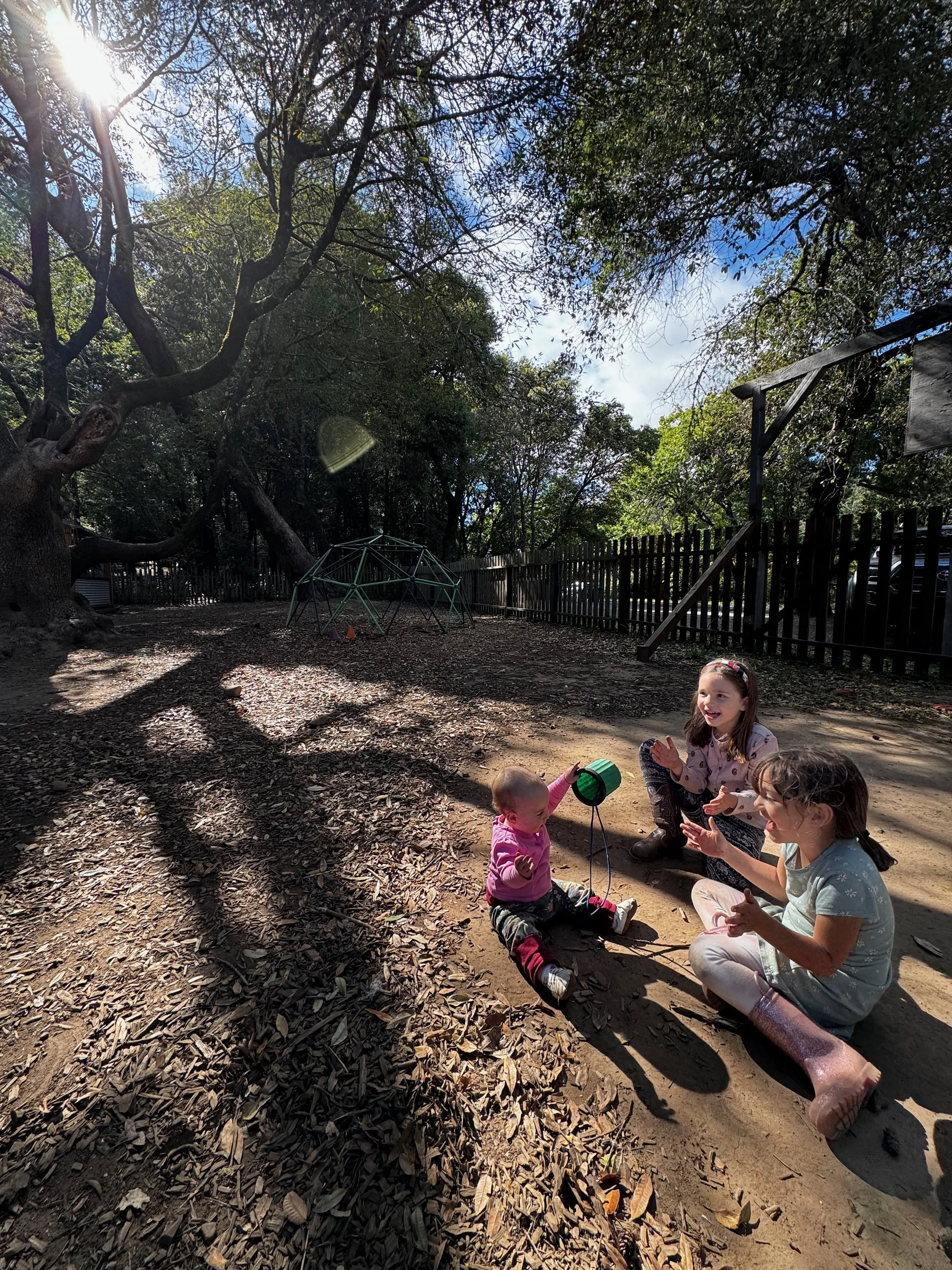 Children sitting on the ground playing with a small green drum in a wooded playground.