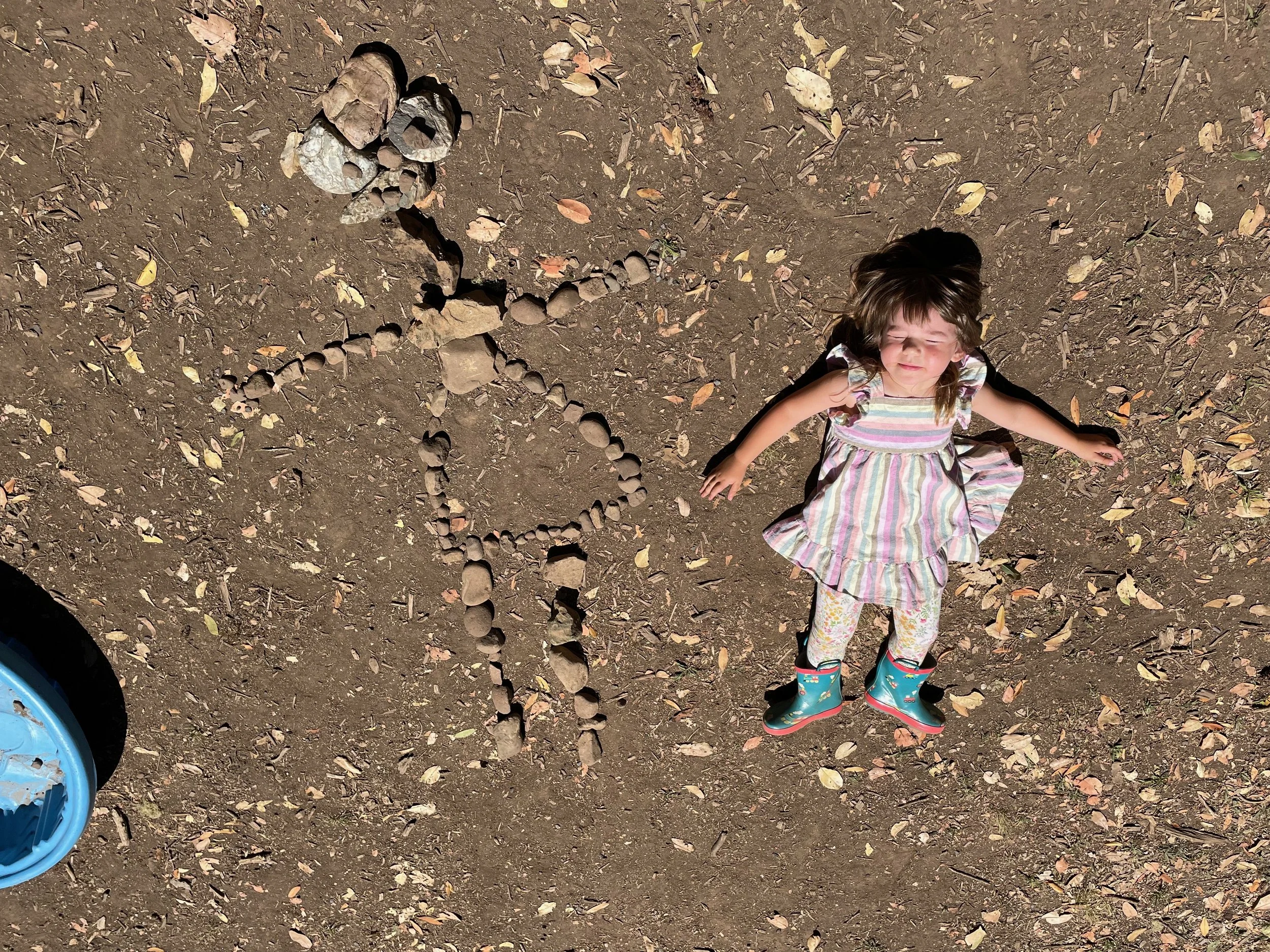 Lying on the ground with her eyes closed, wearing a colorful striped dress and rain boots. Next to her, rocks are arranged to form a butterfly and a flower on dirt ground with scattered leaves.