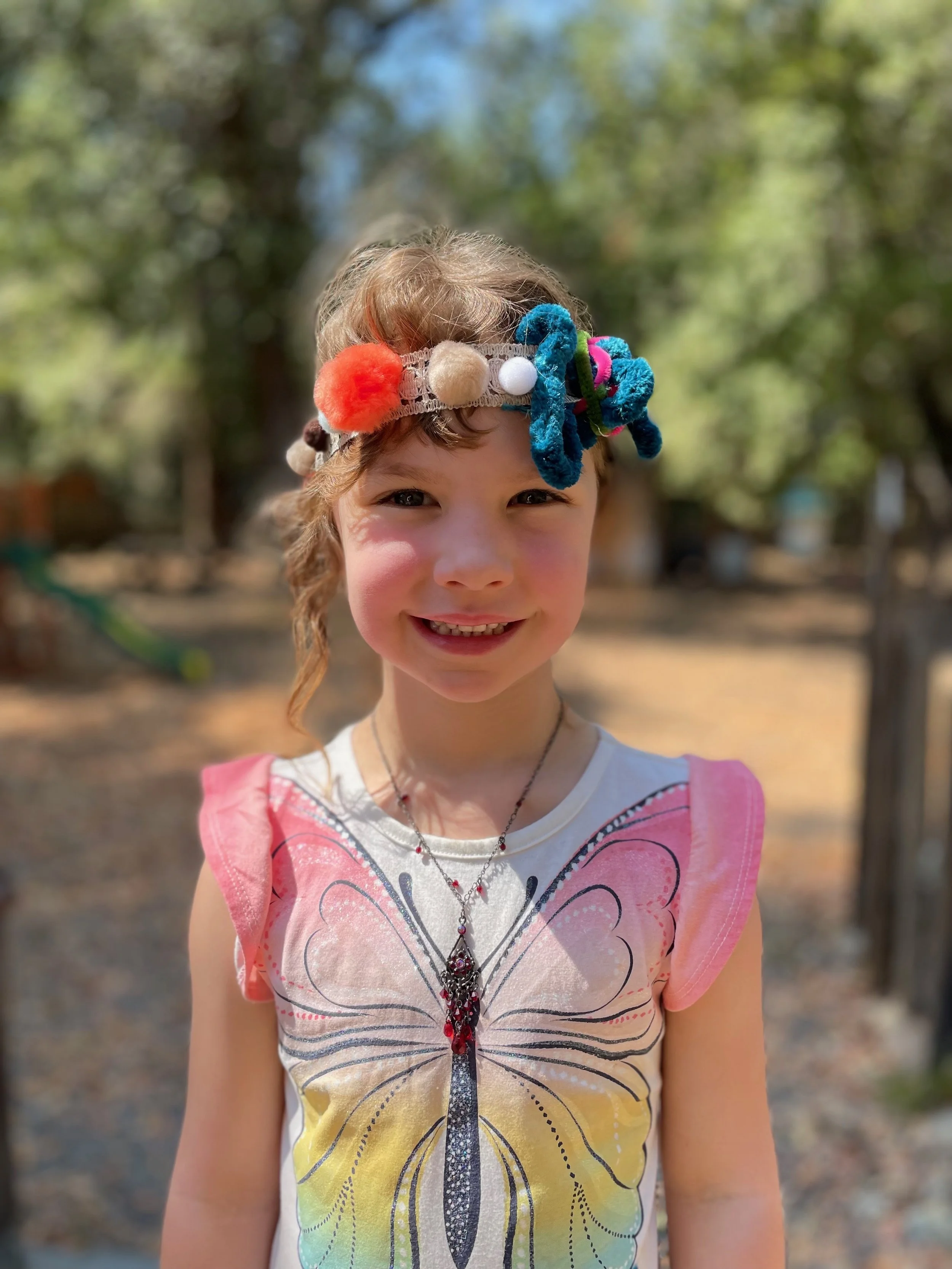 Smiling outdoors, wearing a colorful butterfly print dress and a decorative headband with pom-poms and yarn accessories.