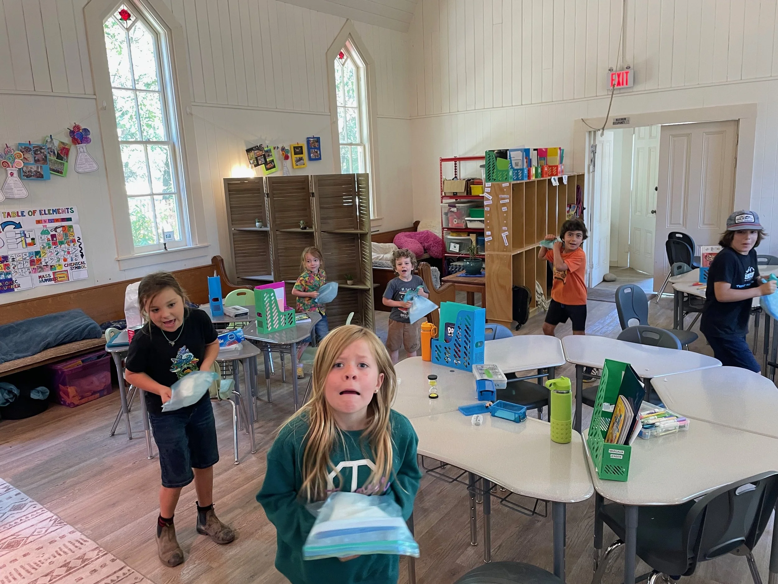 Children in a classroom holding up supplies and engaging in activities, with books, educational posters, and colorful storage bins visible around the room.