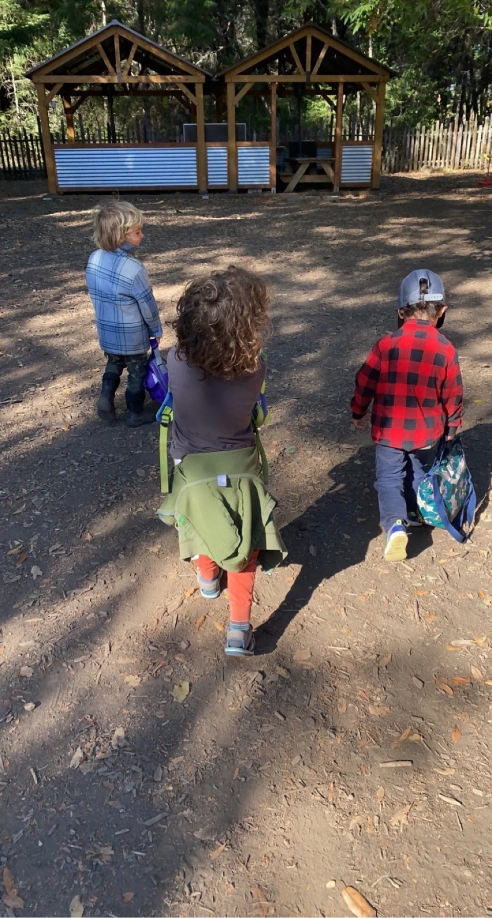 Three children walking away from camera toward a wooden structure with a fence in the background, carrying backpacks, in a dirt outdoor setting with trees.