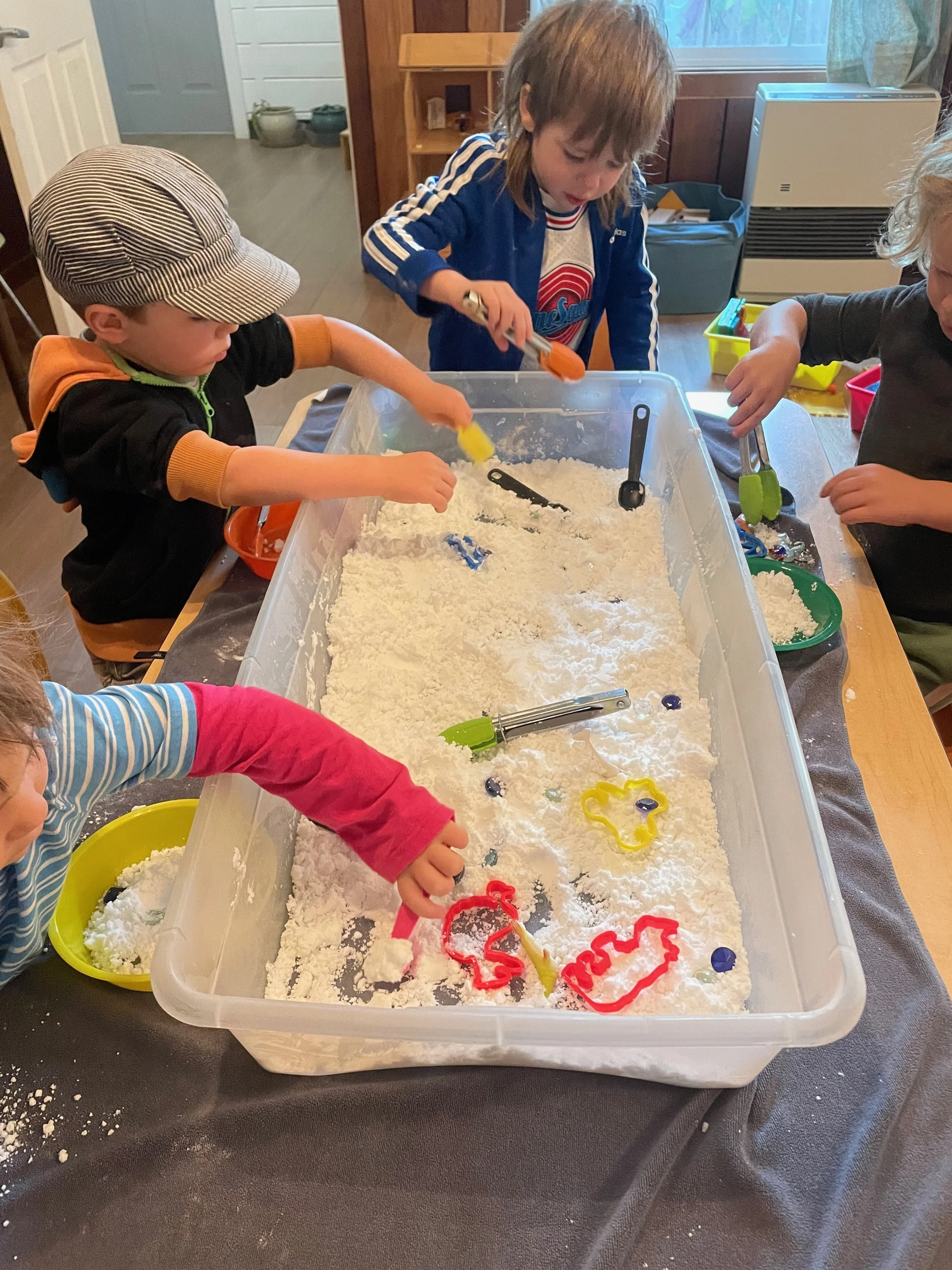 Children playing with kinetic sand and colorful tools at a table in a home setting.