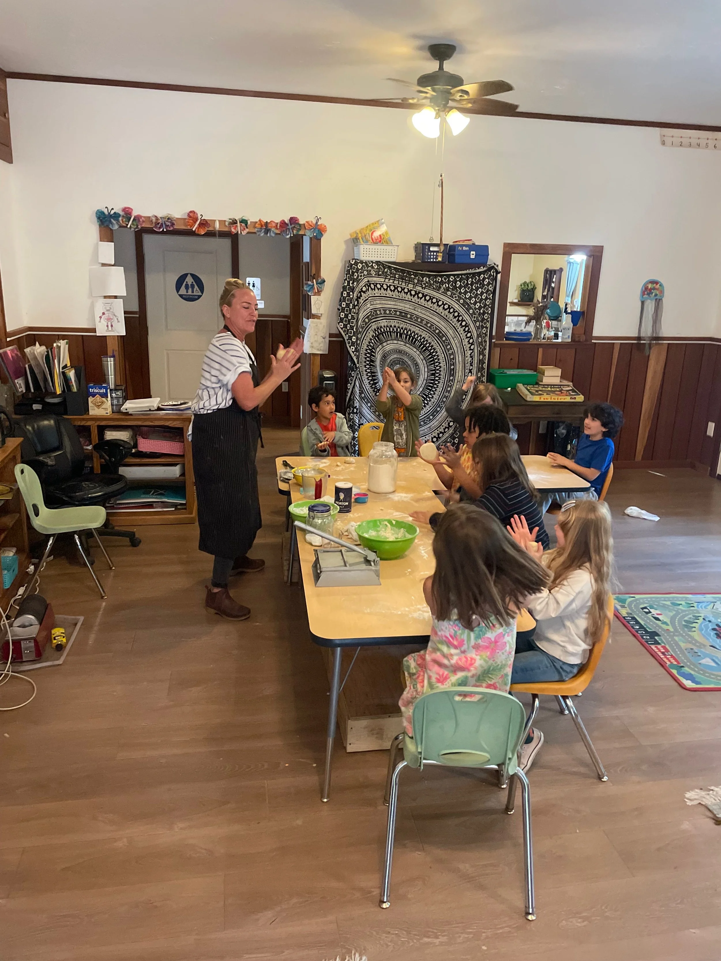 A teacher leading a cooking or baking activity in a classroom with young children seated at a table, some clapping and others raising their hands.