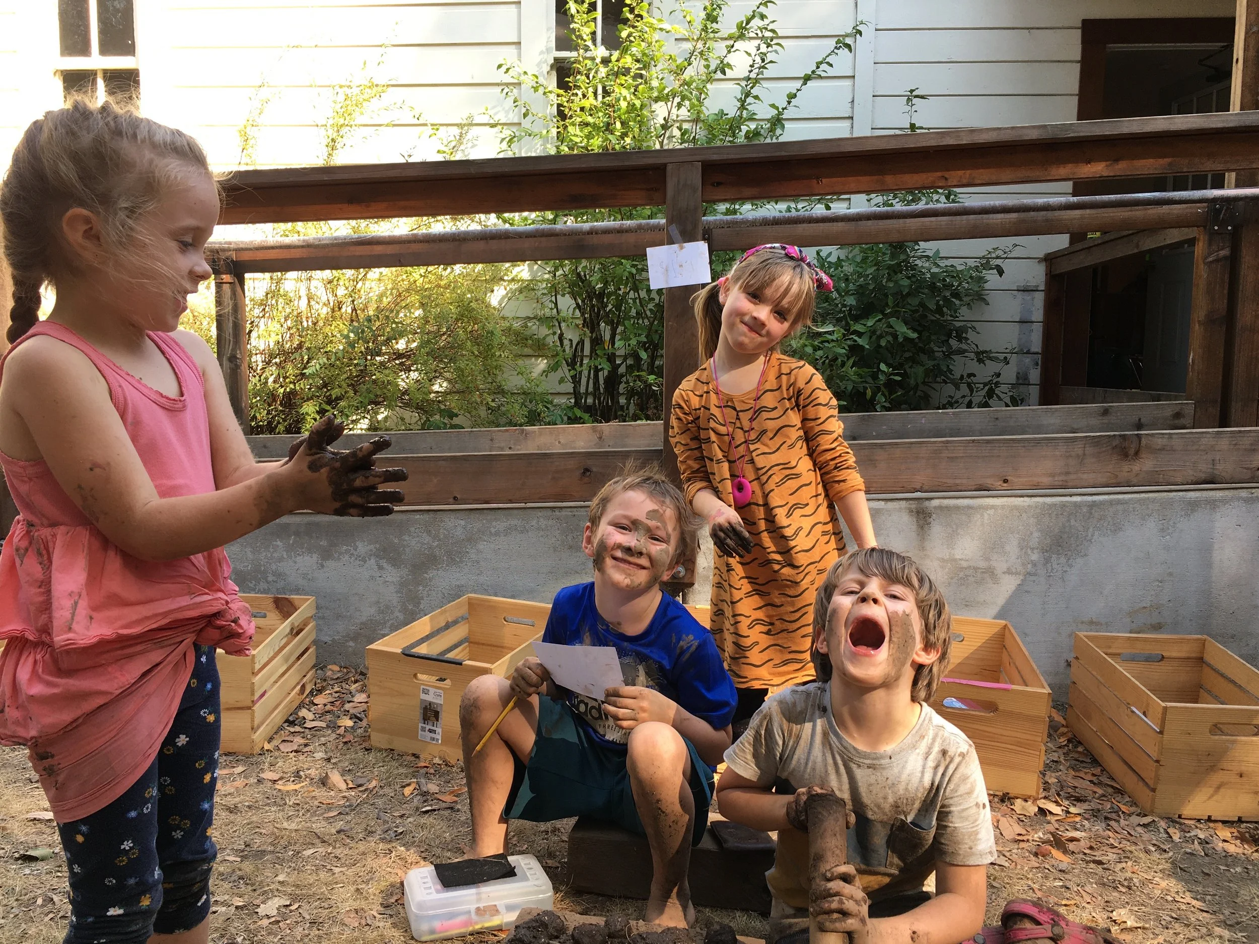 Four children playing outside, covered in mud, with wooden crates behind them and a house in the background.