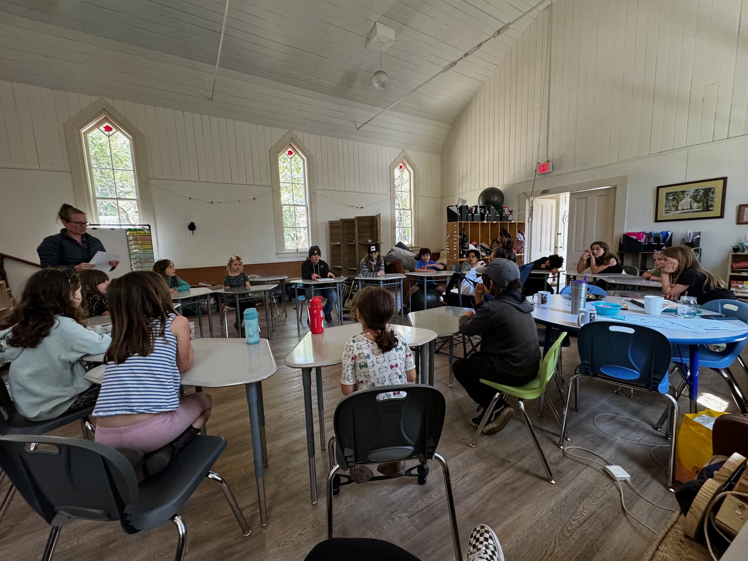 Group of children and a teacher sitting around tables in a classroom, listening to a woman standing at the front reading from papers.