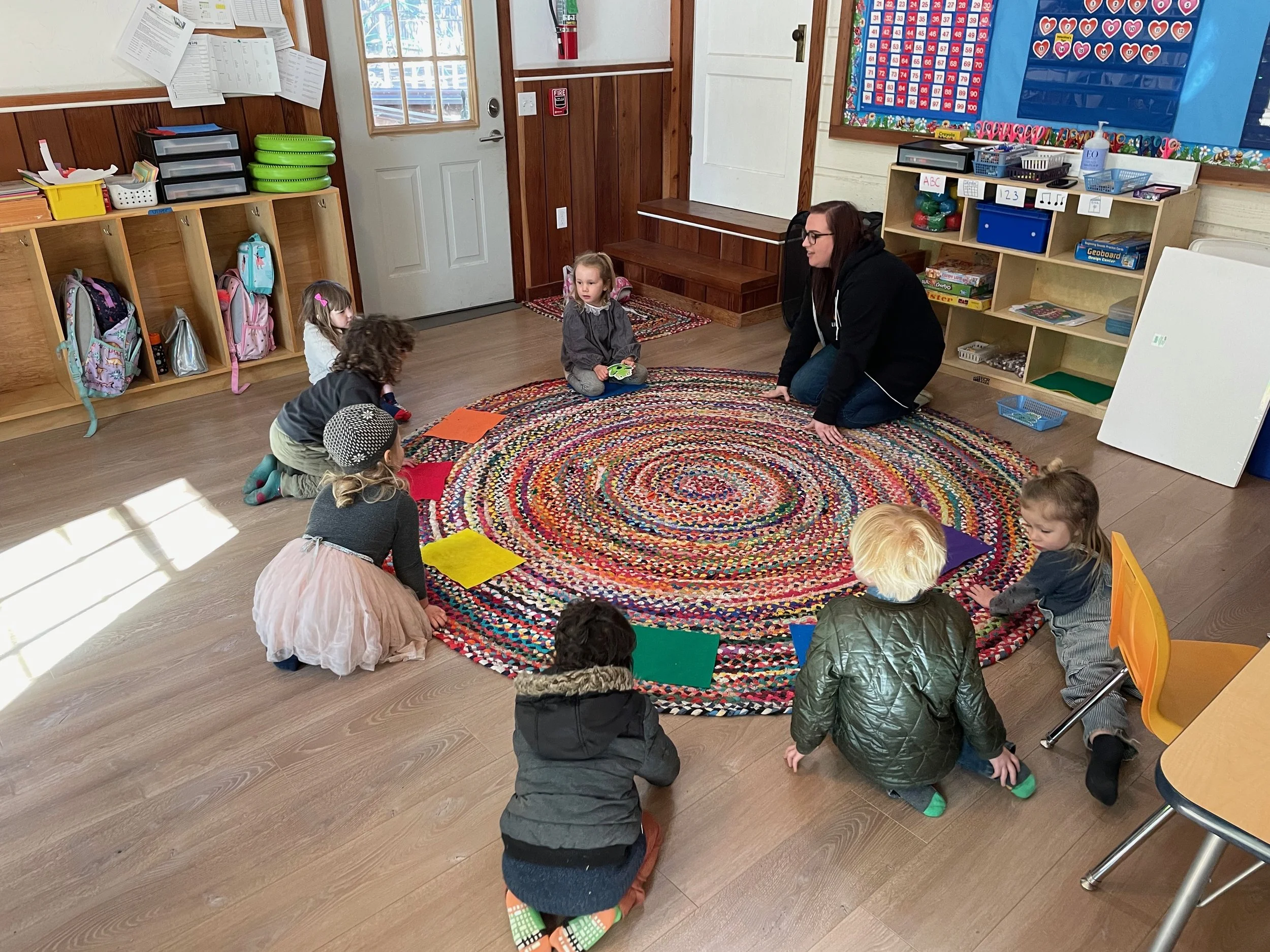A preschool classroom where a teacher is leading a circle time with children sitting around a colorful braided rug.