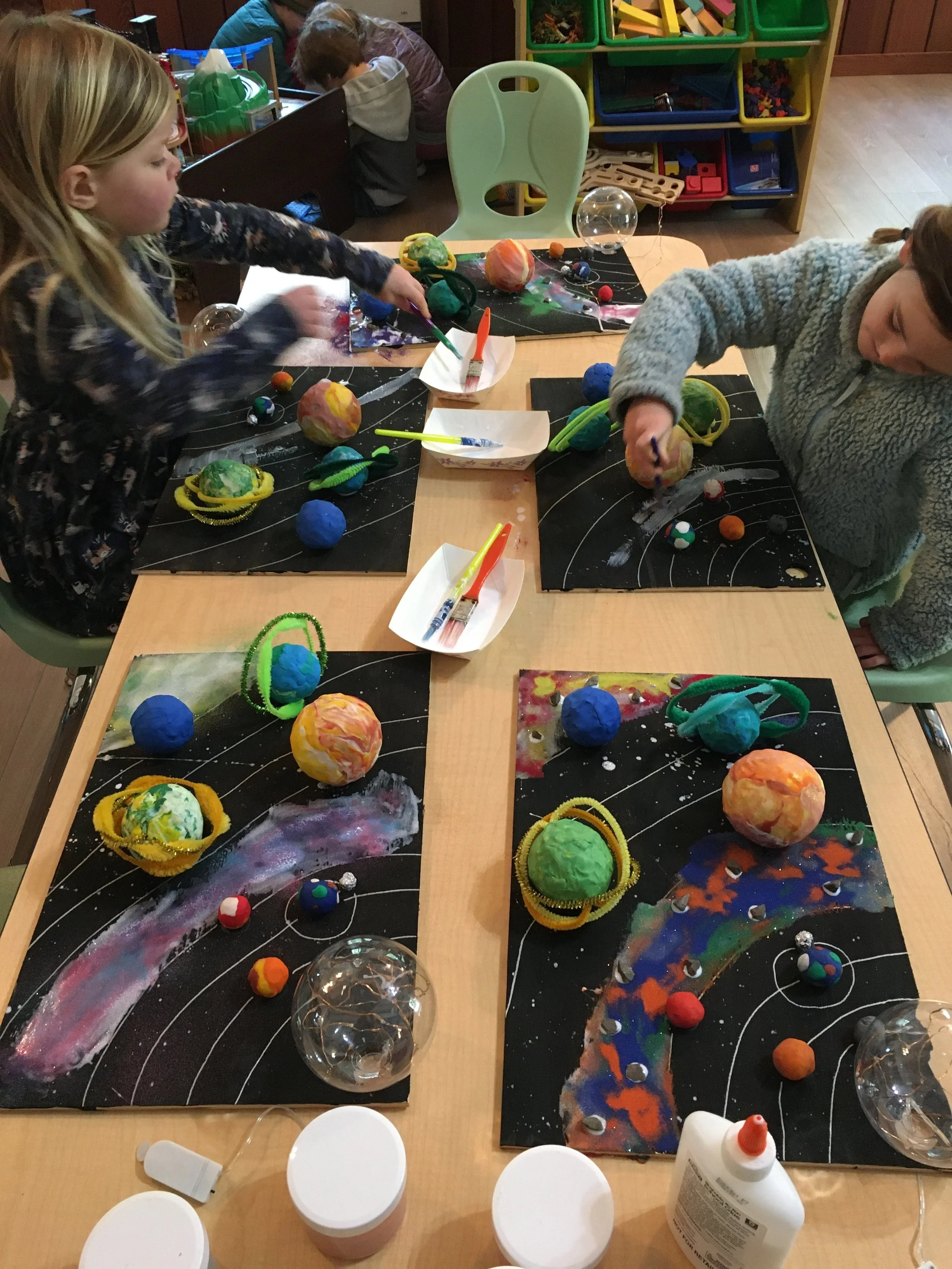 Children creating space-themed art projects with foam balls, pipe cleaners, and paint at a craft table.