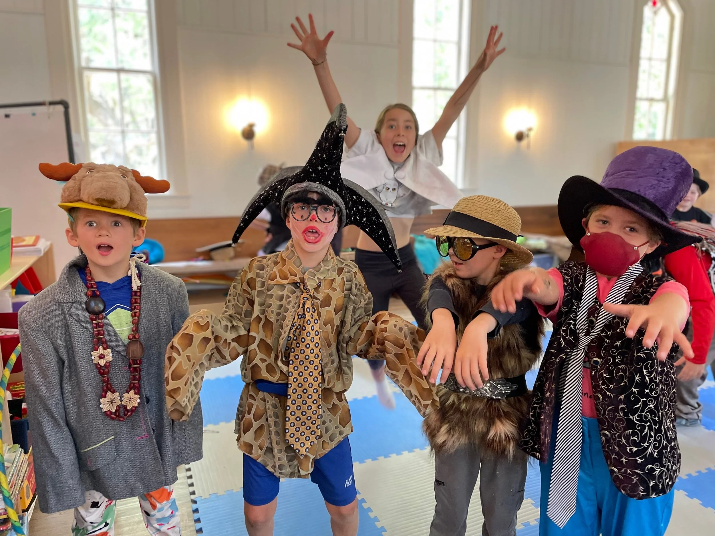 Dressed in costumes, including a boy with a moose hat, a girl with a witch hat and glasses, a girl with a straw hat and sunglasses, a girl with a Wizard hat, and a girl with a starry hat and red mask, posing playfully indoors.
