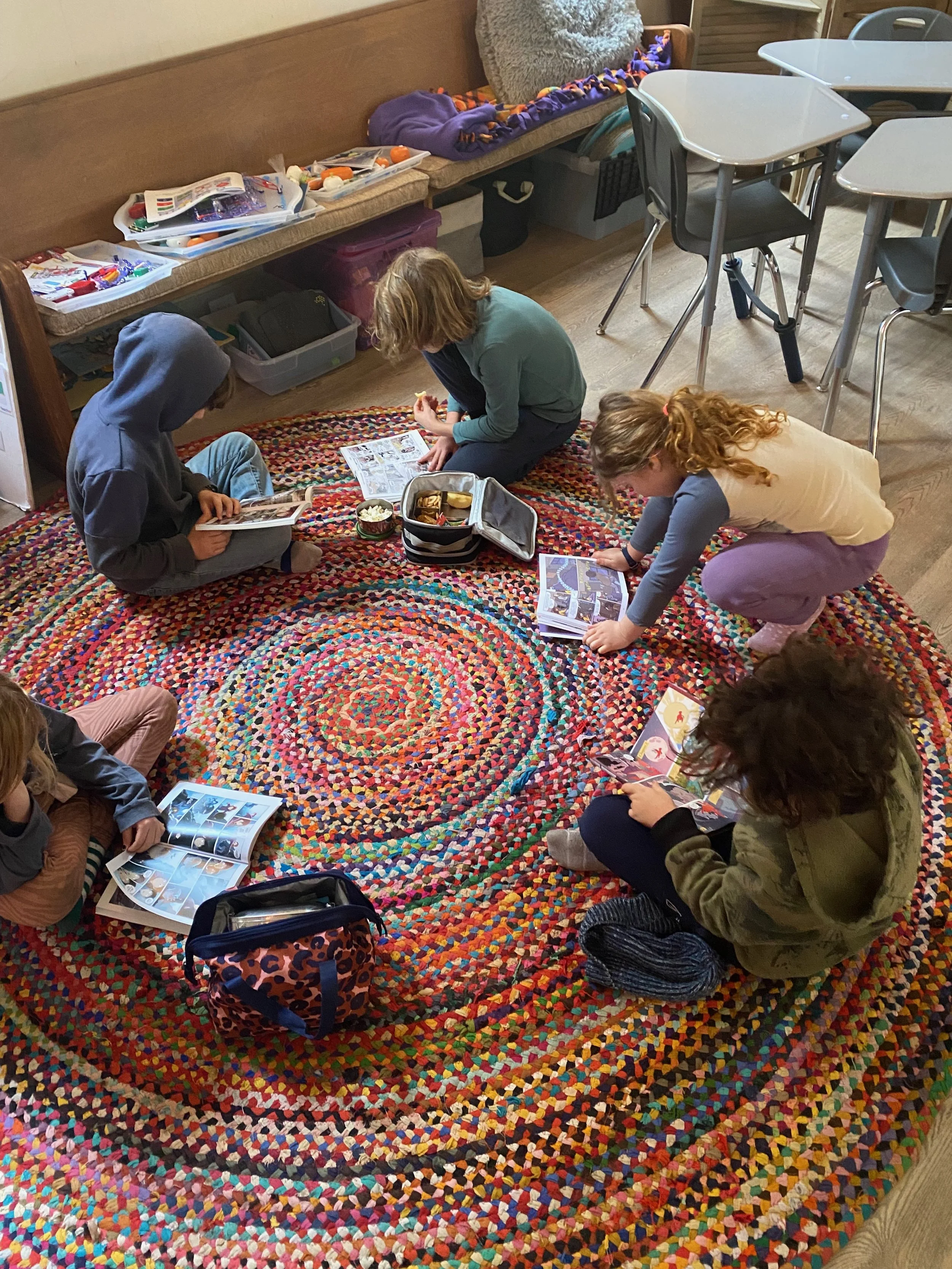 Five children sitting on a colorful, circular braided rug, looking at comic books and magazines.