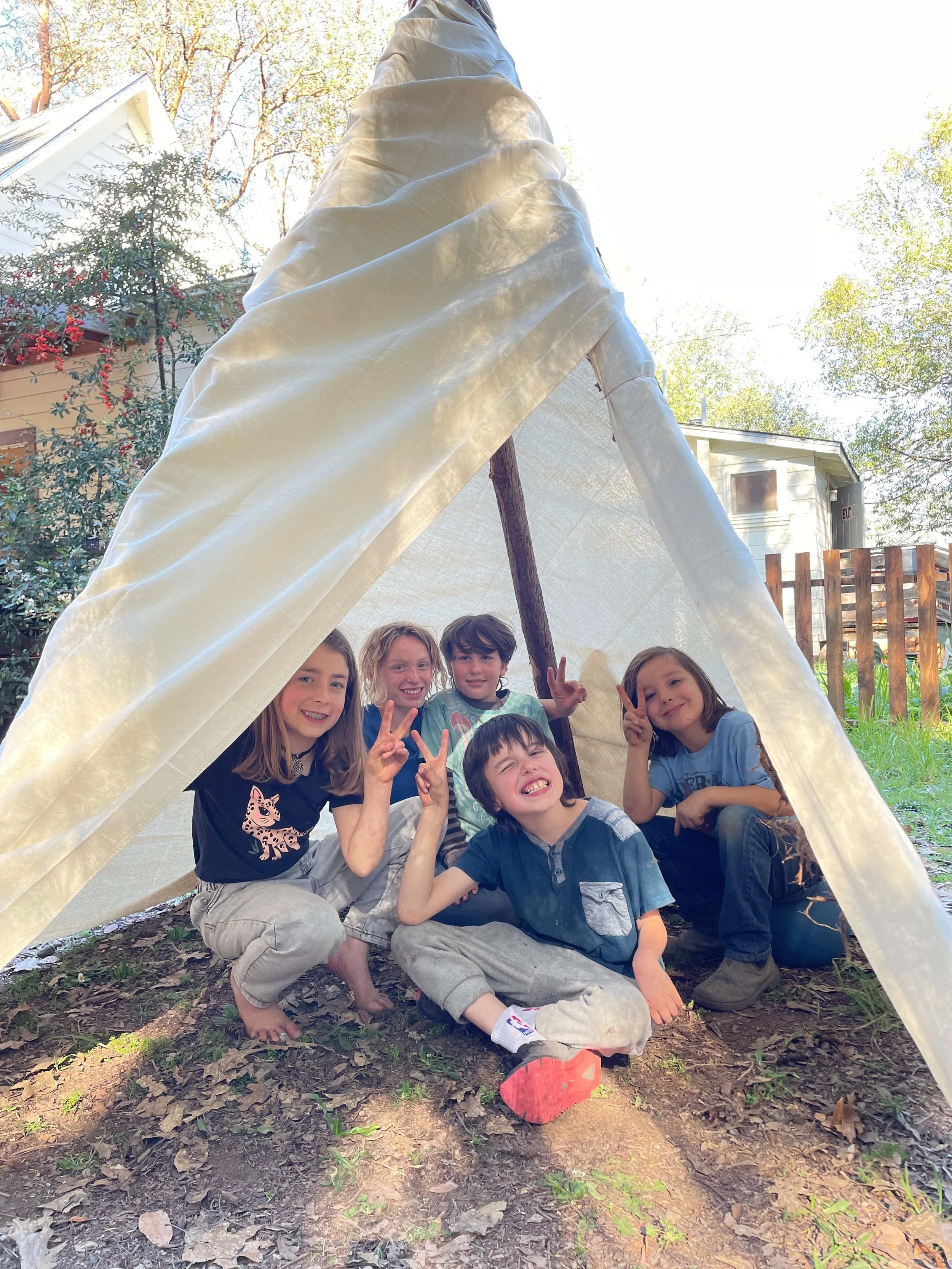 Five children inside a small tent outdoors, smiling and making peace signs, with trees and houses in the background.