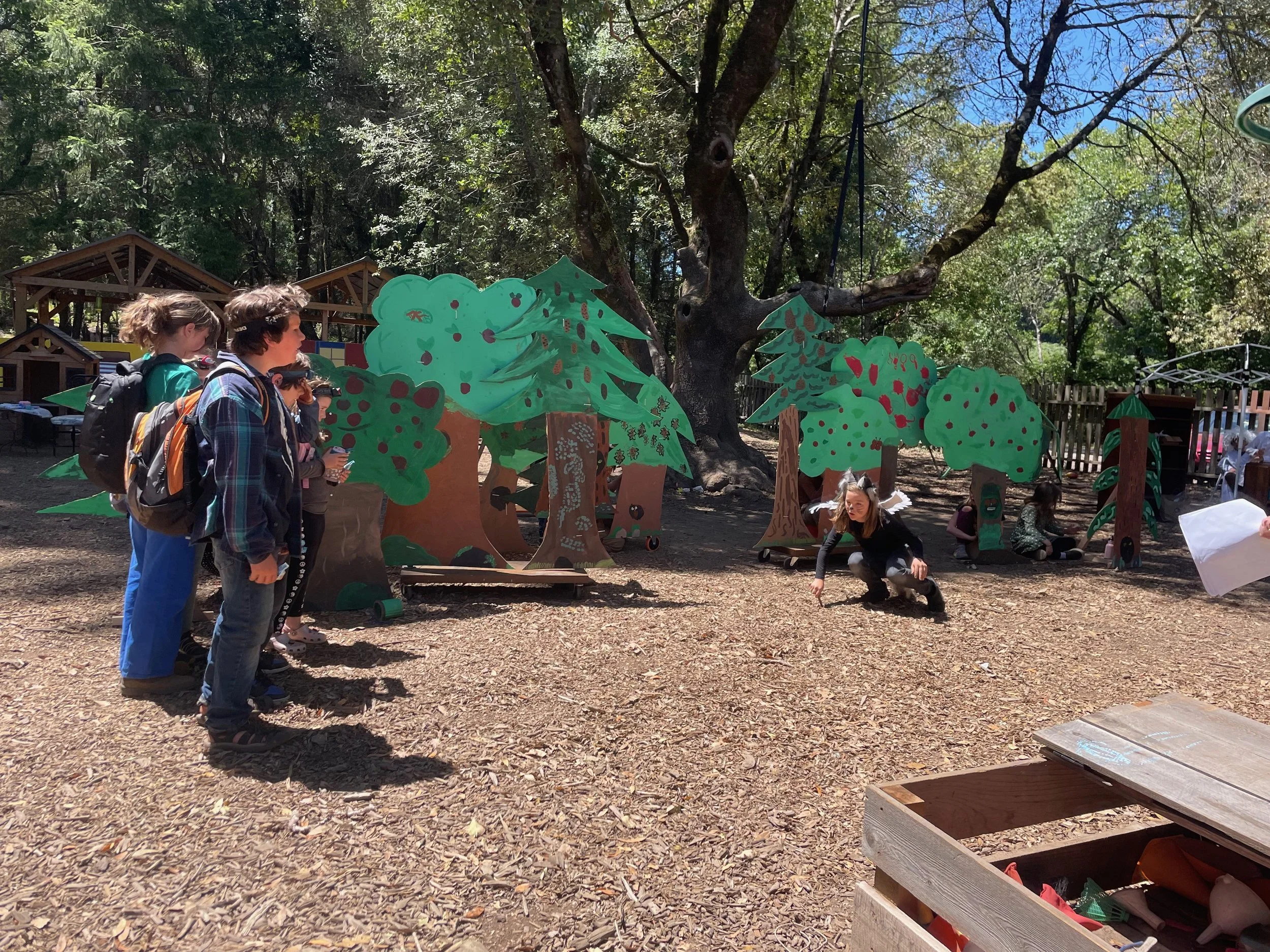 Group of children in costume and others observing along cardboard trees with painted green leaves and red berries, set outdoors under trees on a sunny day.