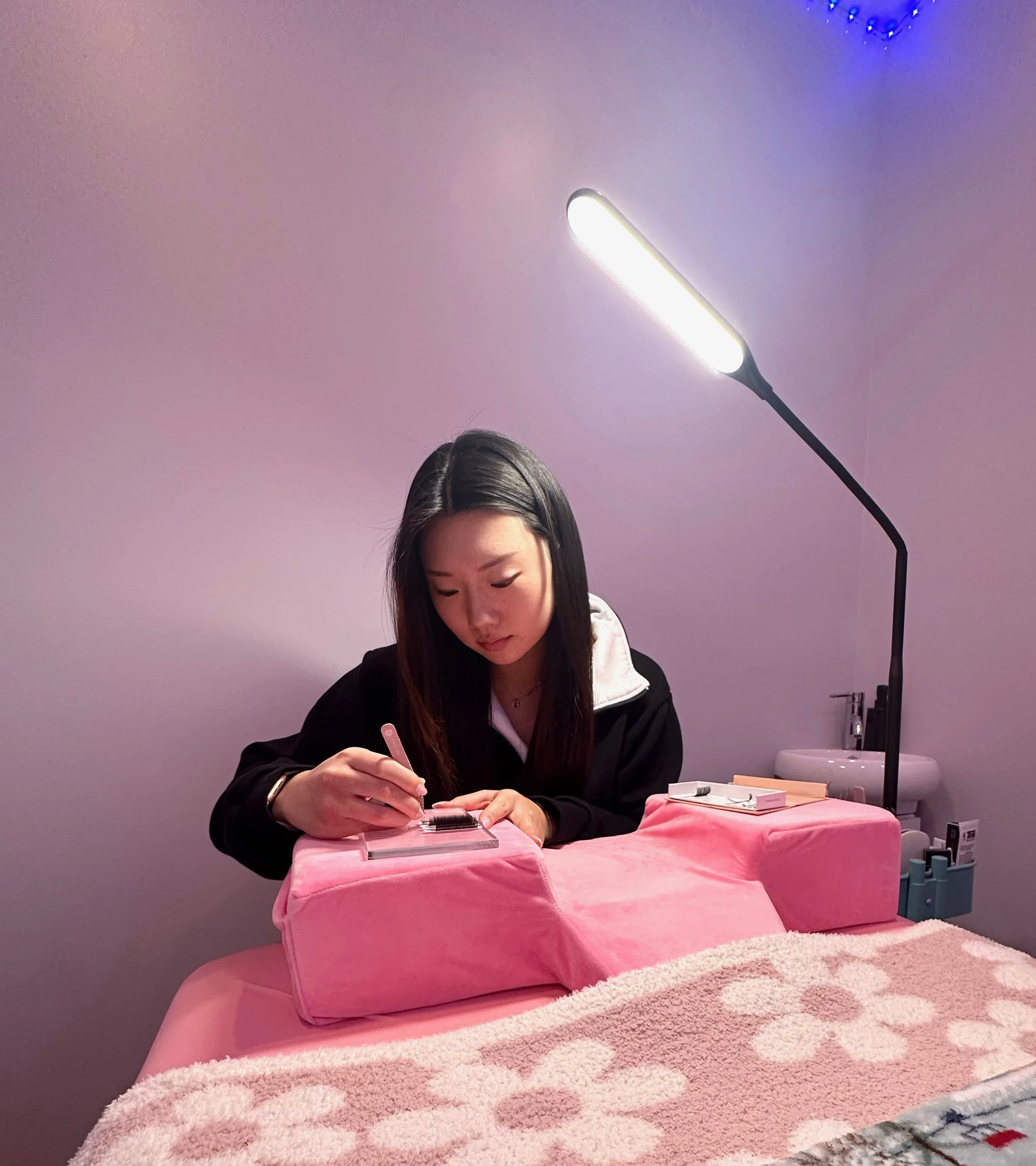 Woman working at a pink-covered table with a LED lamp, focusing on small objects in a nail or beauty salon setting.