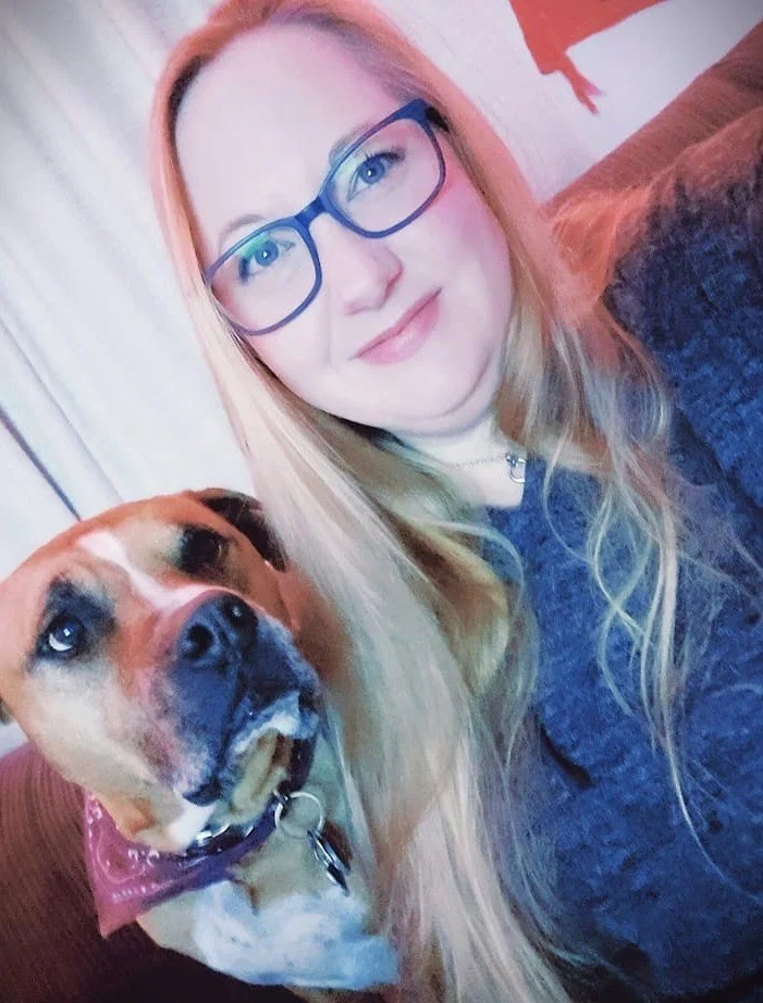 A woman with long blonde hair, wearing glasses, and a black top, sitting next to a large brown and white dog with a pink bandana, on a brown couch.