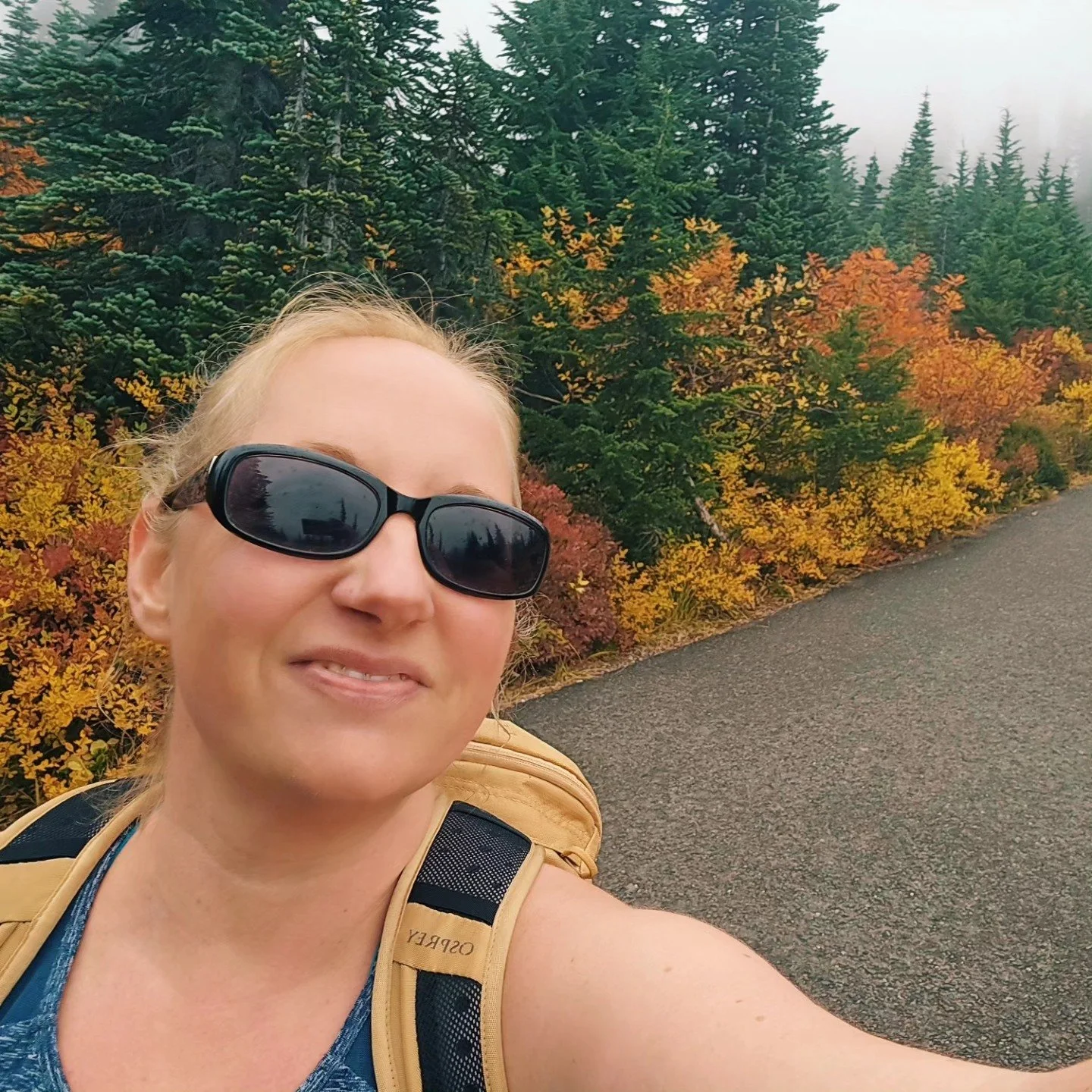 A woman with blonde hair wearing sunglasses and a beige backpack, smiling in front of autumn-colored trees and green pine trees on a cloudy day.