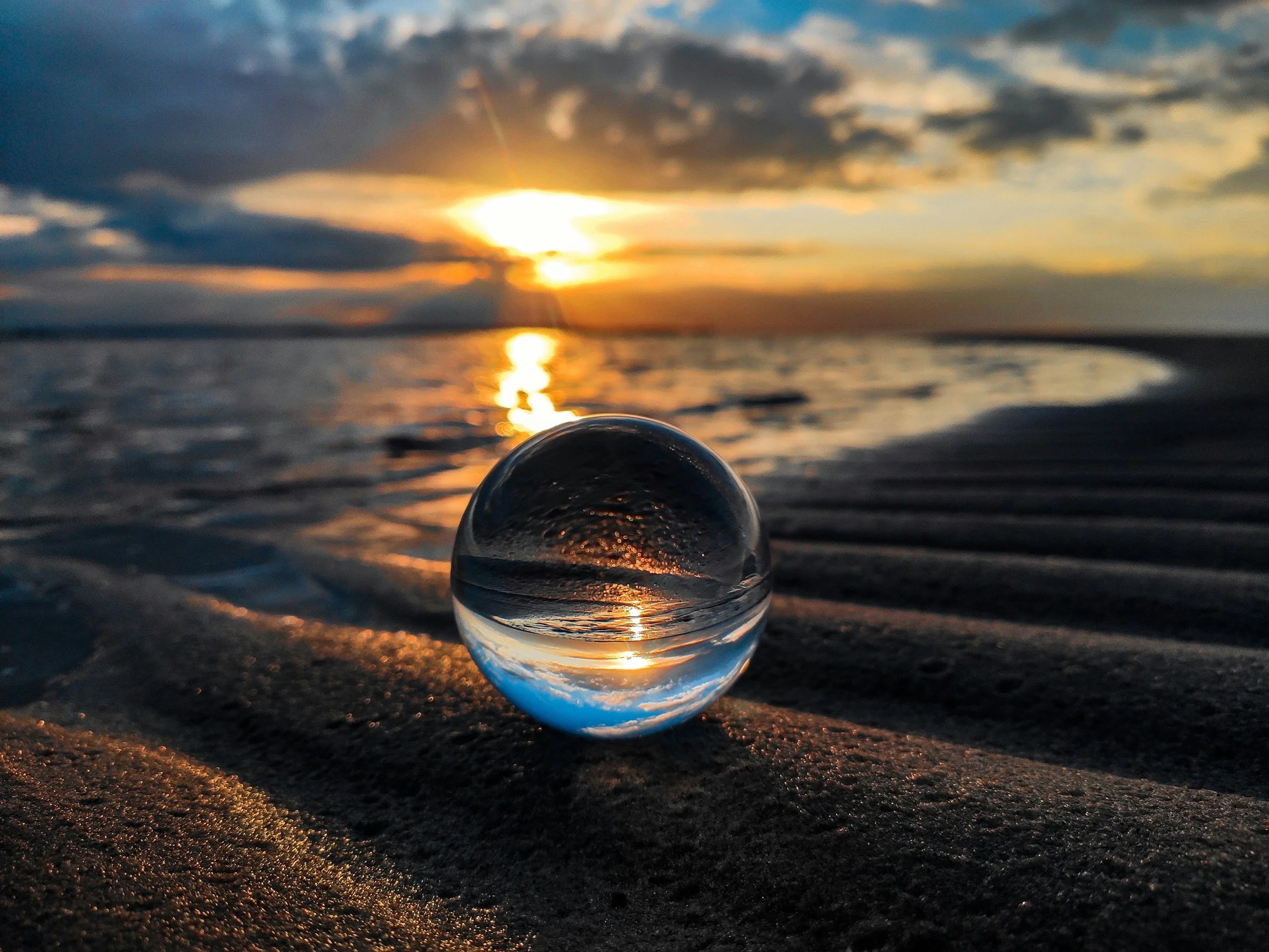 A glass sphere on sand at the beach reflects the sunset sky, with clouds and the sun visible on the horizon, and the sky displays warm colors.