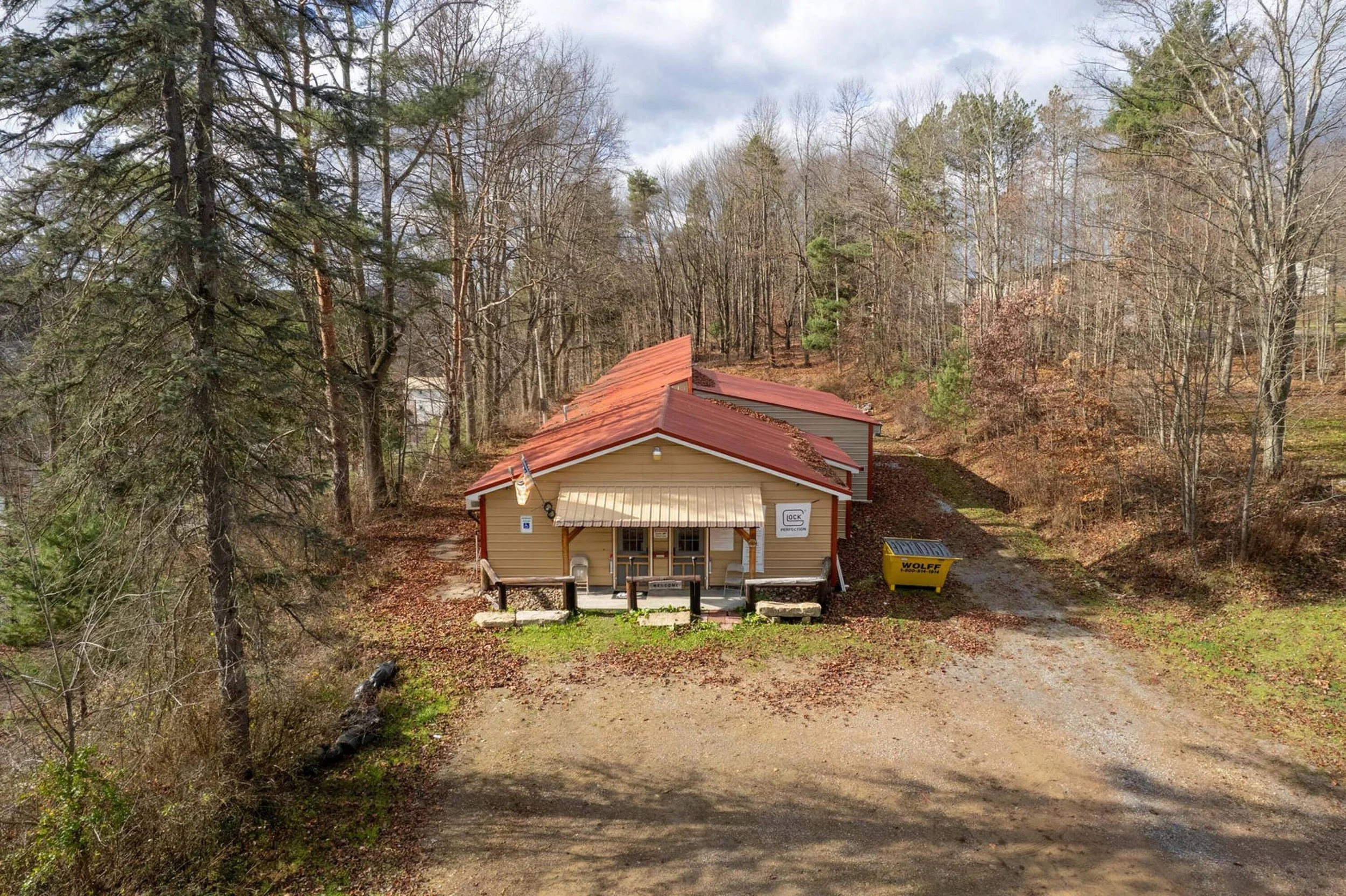 A small building with a red metal roof, beige siding, and a porch with chairs, surrounded by trees and a gravel driveway.