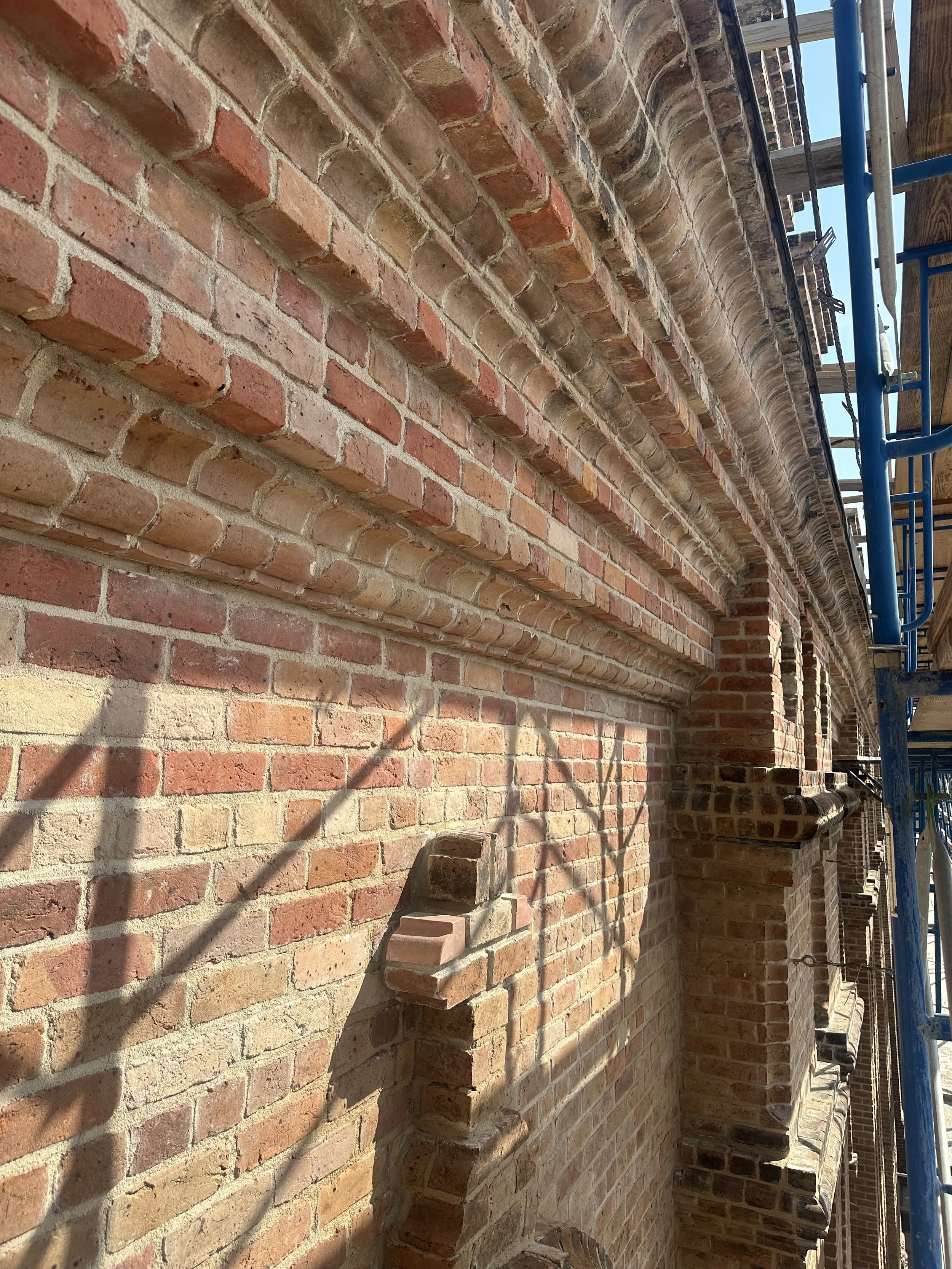 Brick exterior wall of a building under construction with scaffolding, shadows of scaffolding cast on the wall, showing brickwork and some architectural details.