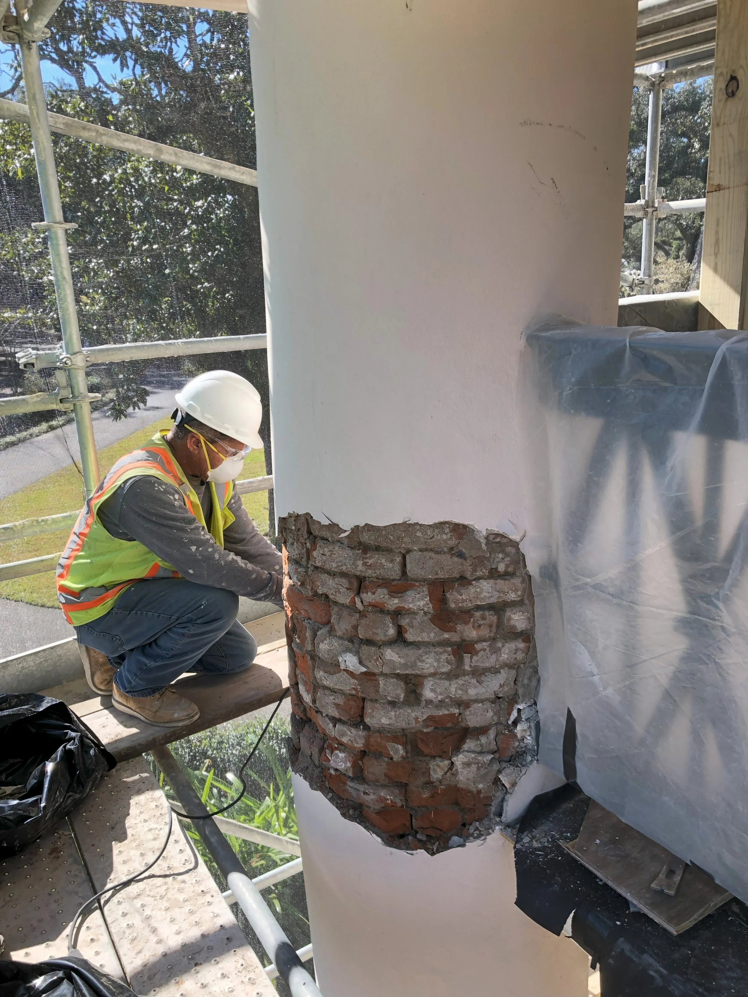 A construction worker wearing a white helmet, safety goggles, face mask, yellow safety vest, gray long-sleeve shirt, and jeans is squatting on a wooden platform. The worker is working on a brick column attached to a white wall, with construction scaffolding and trees visible in the background.