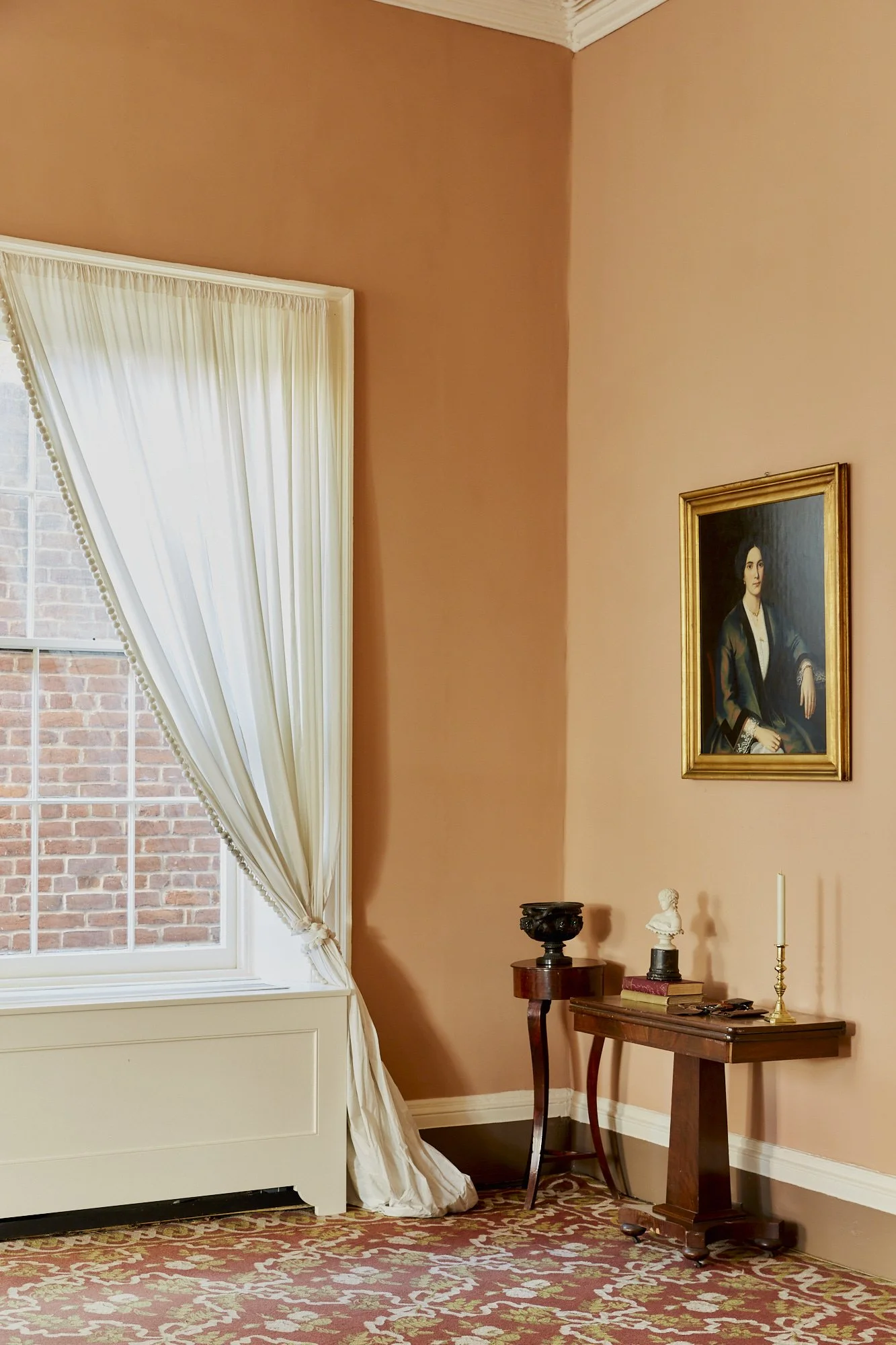 A corner of a room with a window with white curtains, a portrait painting on the wall, a small wooden table with decorative objects, and a patterned carpet.