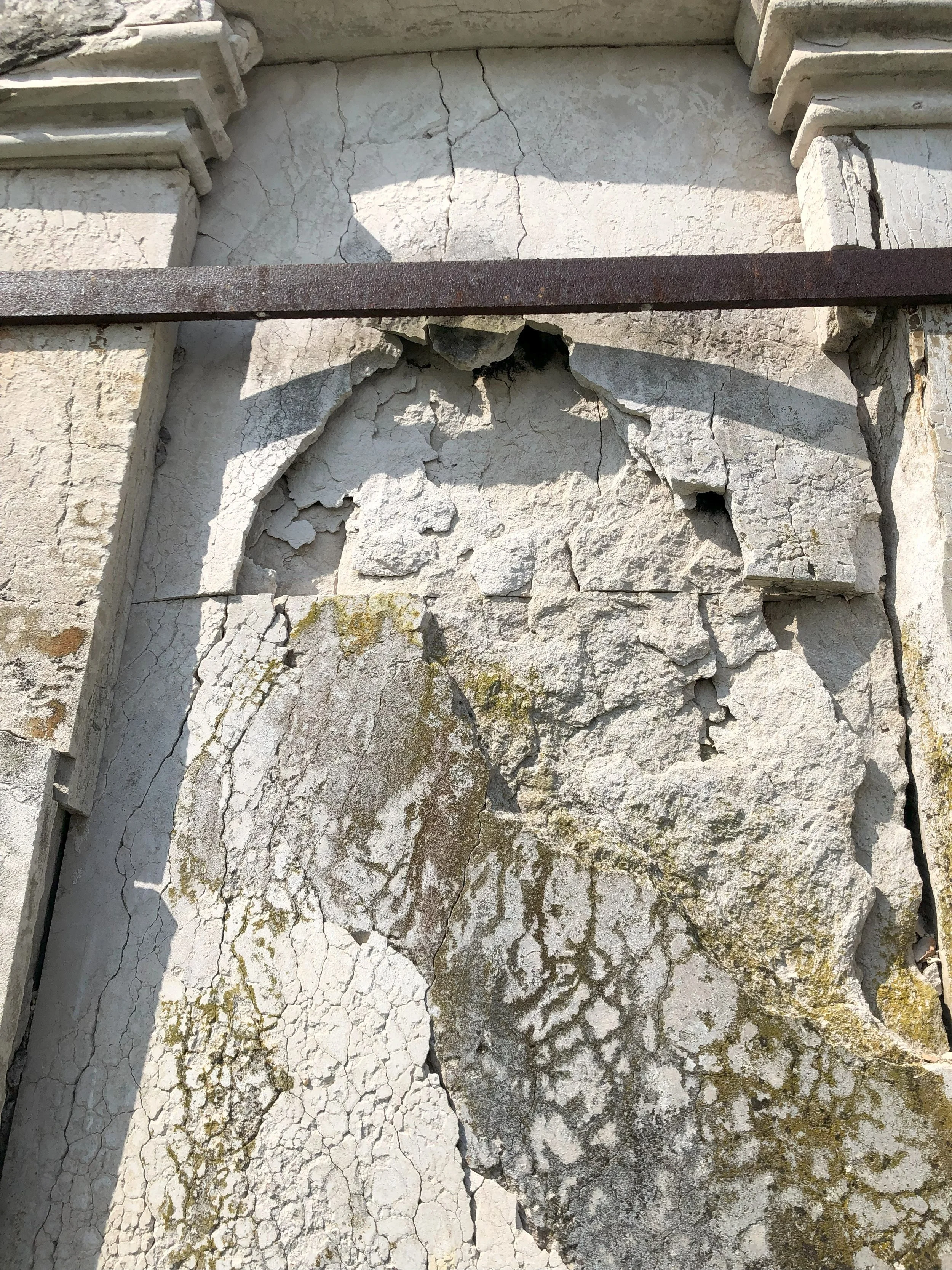 Close-up of crumbling, cracked white stone or plaster wall with moss growth and rusted metal bar across top.