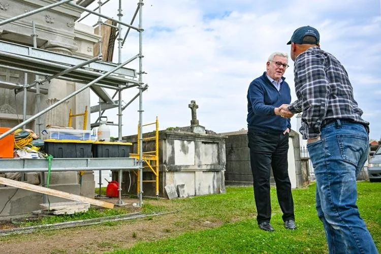 Two men standing outside near a building under construction, with scaffolding on the left, engaging in a handshake or exchange.