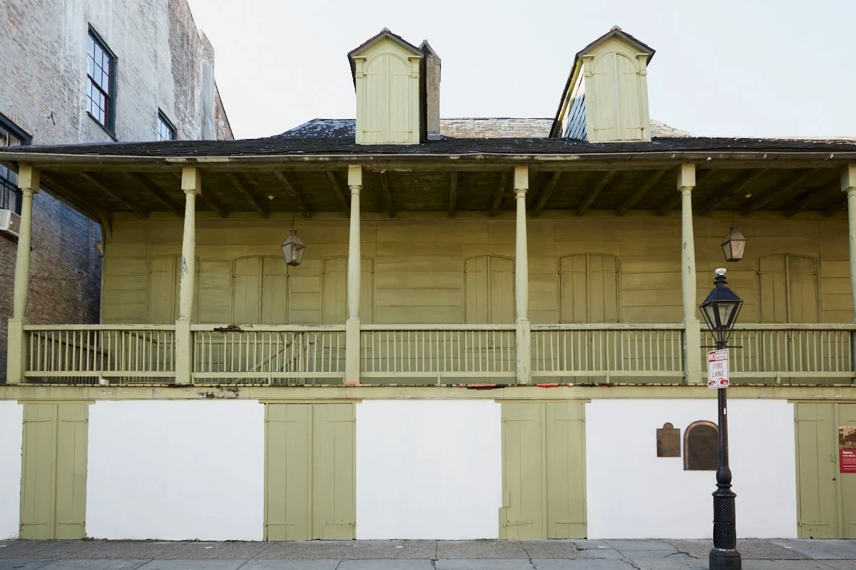 A two-story wooden building with a balcony, painted in pale yellow, featuring closed shutters on the upper windows, hanging lanterns, and a street lamp in front. A sign indicates no fire lane parking.