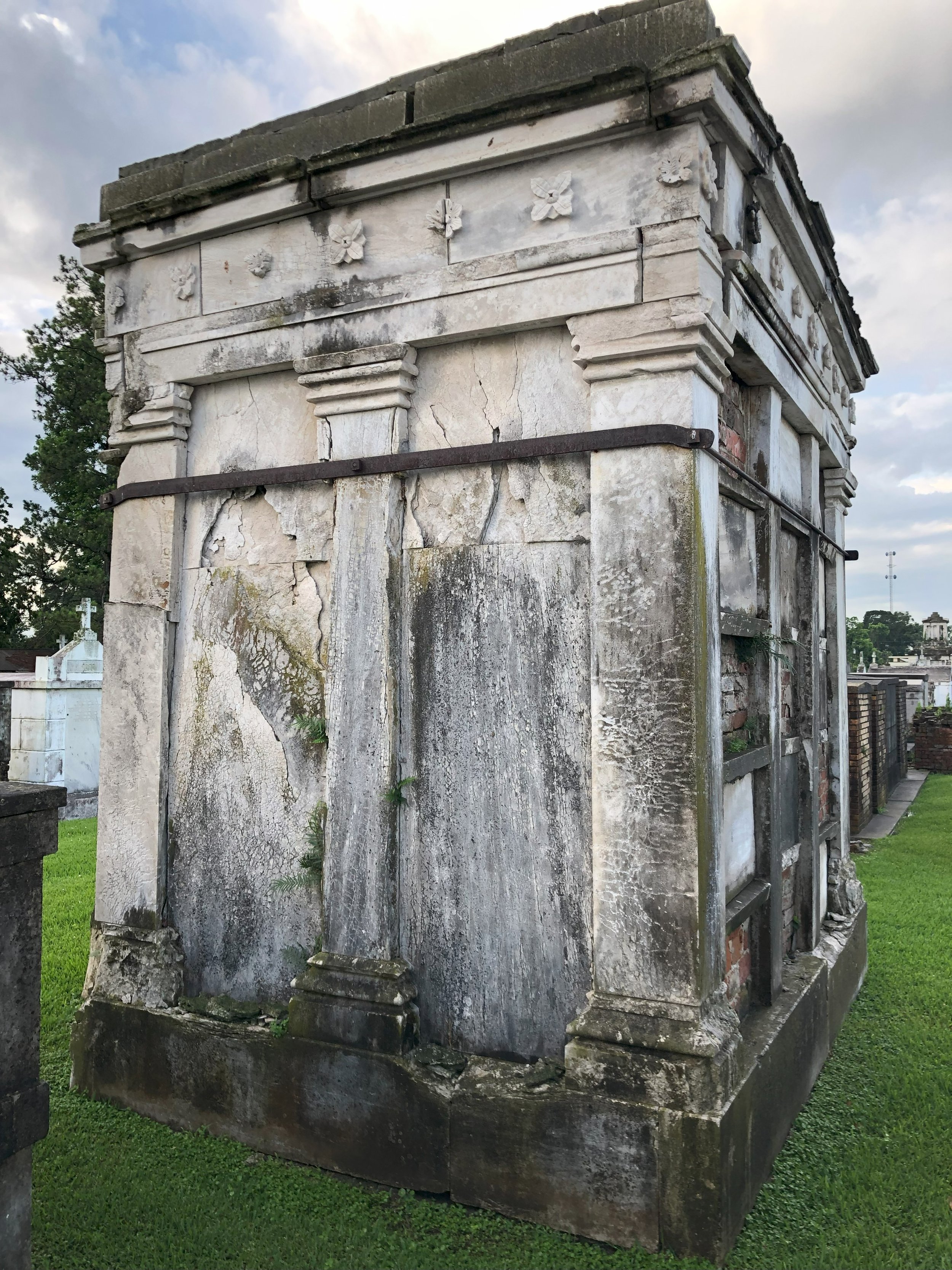 Old, weathered mausoleum with cracked stone and overgrown plants, situated in a grassy cemetery.