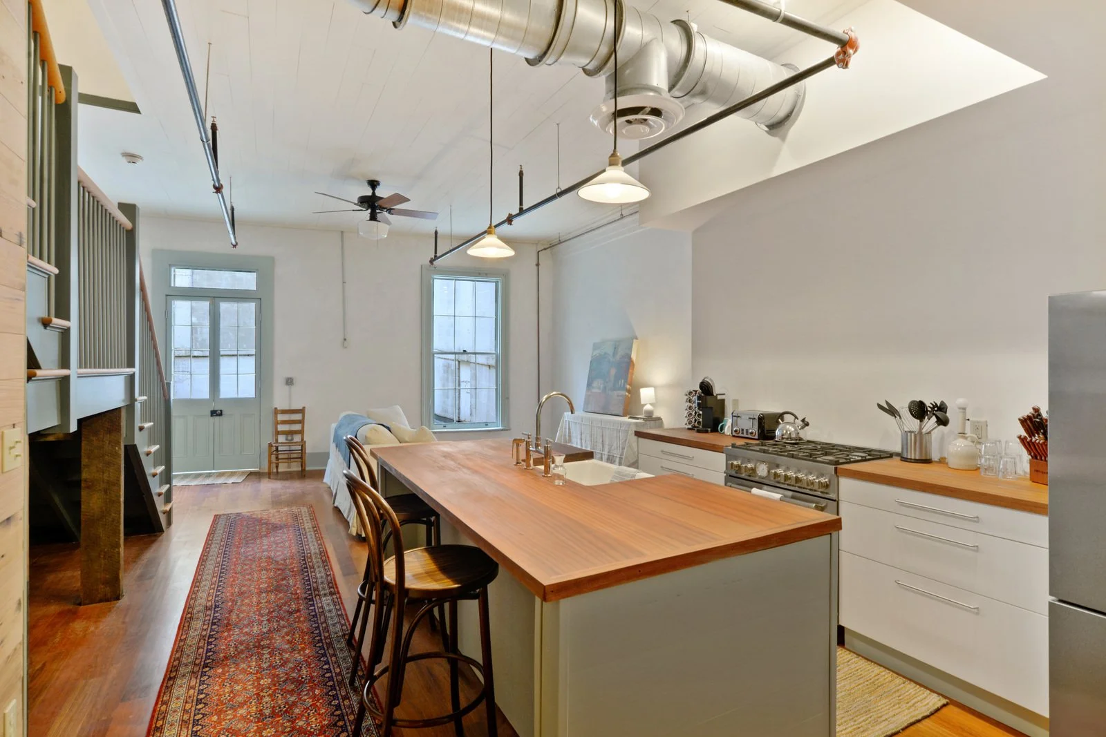 Open-concept kitchen and living area with wooden flooring, white walls, large windows, and an exposed industrial-style ceiling with ducts. The kitchen features a wooden countertop island, white cabinets, and stainless steel appliances. There is a small rug on the floor, and a hallway leading to a door and windows at the back.