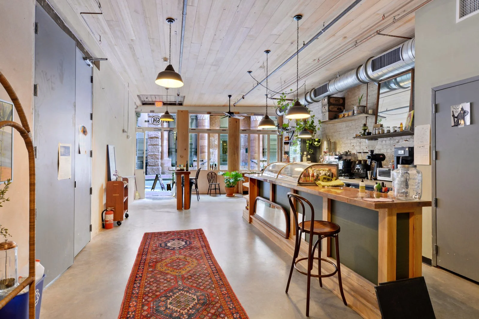 Interior of a cozy coffee shop with a long wooden counter, hanging pendant lights, a display case for baked goods, and a few chairs and tables near the large front windows.