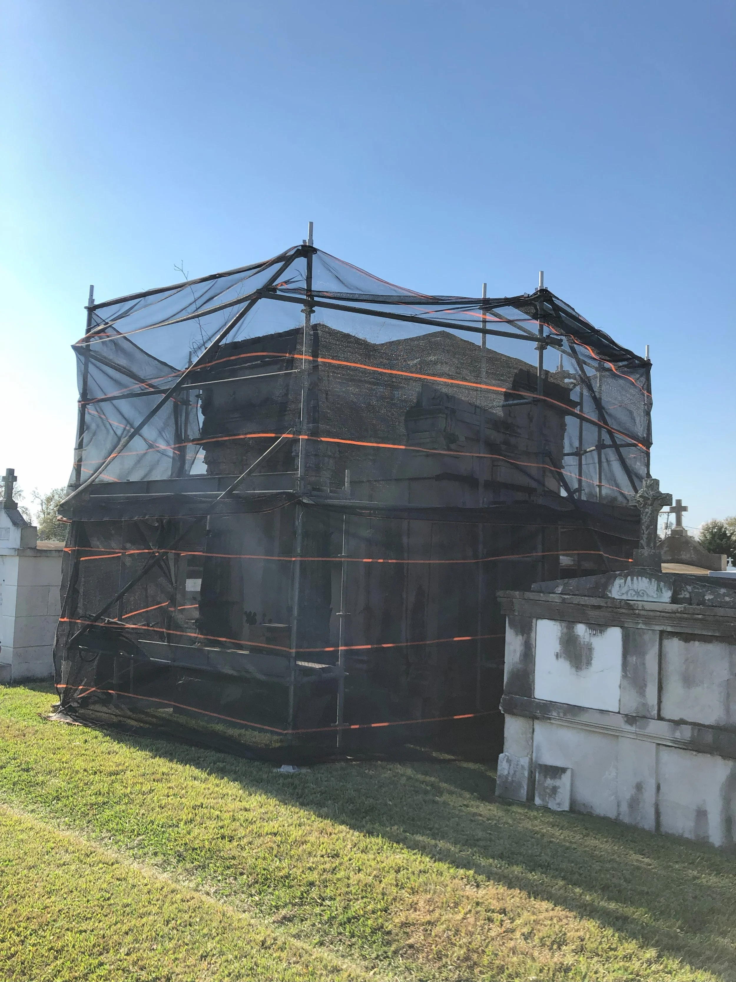 A mausoleum surrounded by black protective netting for restoration or maintenance work, situated in a cemetery with grass and other tombstones visible.