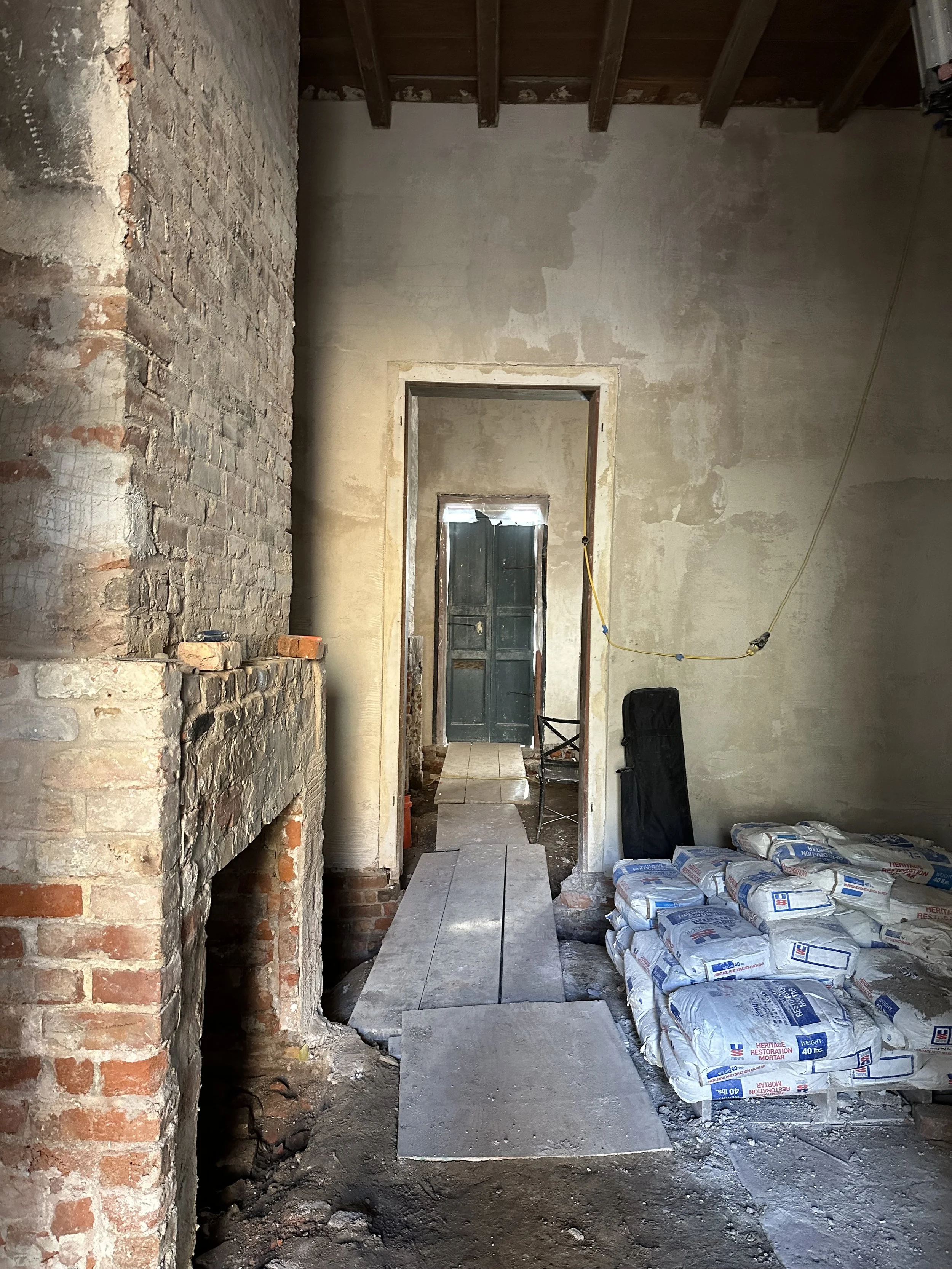 Interior of a building under renovation with exposed brick, unfinished walls, and construction materials such as cement bags and wooden planks on the floor, leading towards a doorway with a view of a partially painted green door.