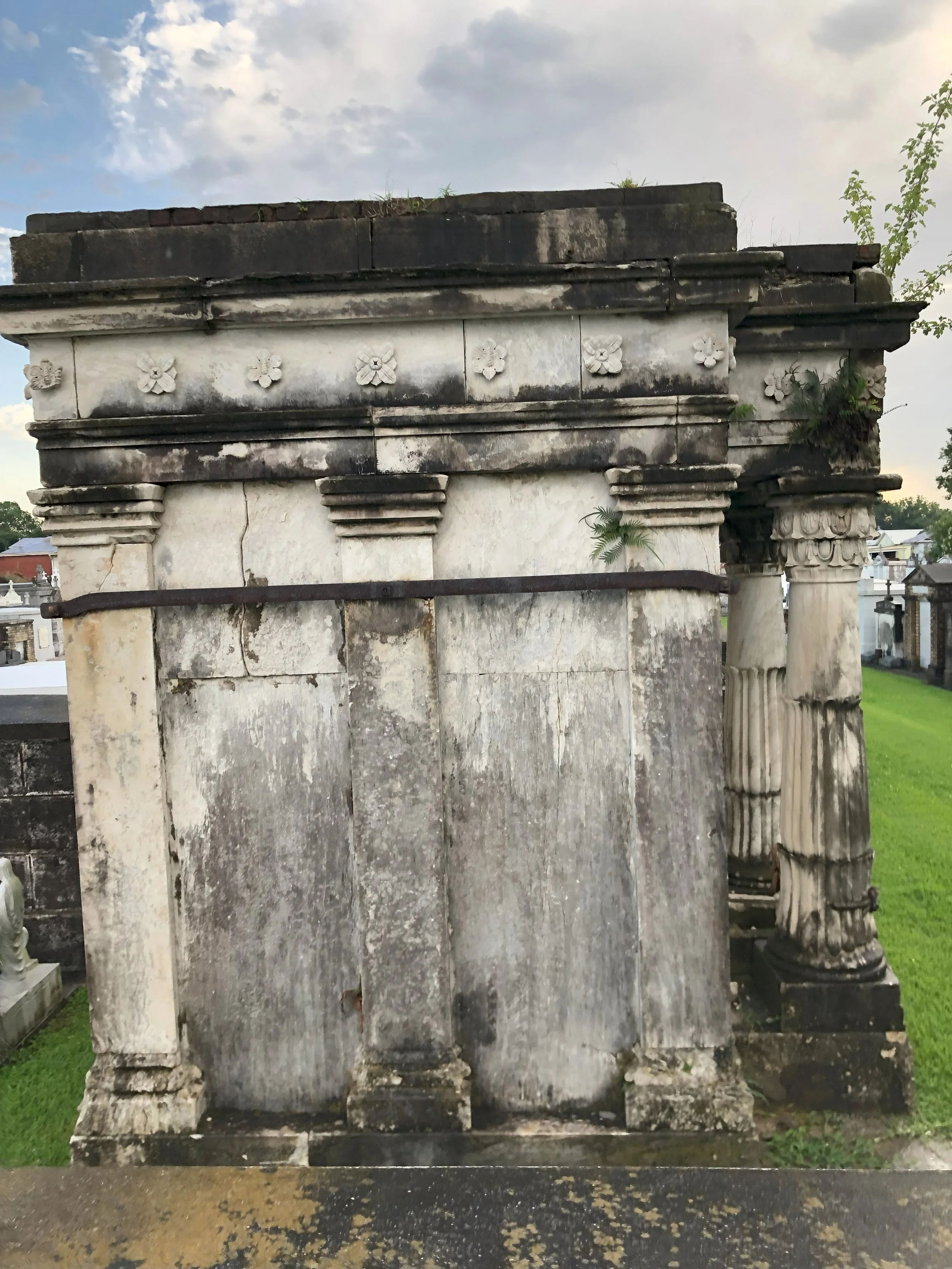 An old, weathered stone monument with columns and decorative flower motifs on the upper part, situated in a cemetery with a grassy area in the background.