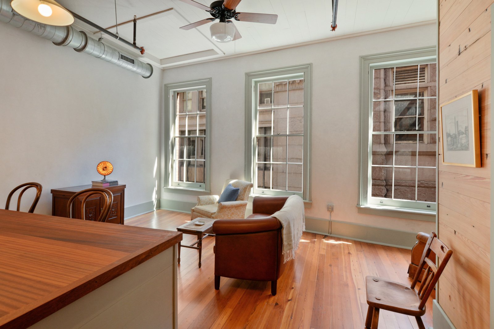 Interior view of a bright living room with three large windows, hardwood floor, a ceiling fan, and wooden accent wall. Furnished with two armchairs, a leather chair, a small side table, and an antique wooden sideboard with a small decorative lamp.
