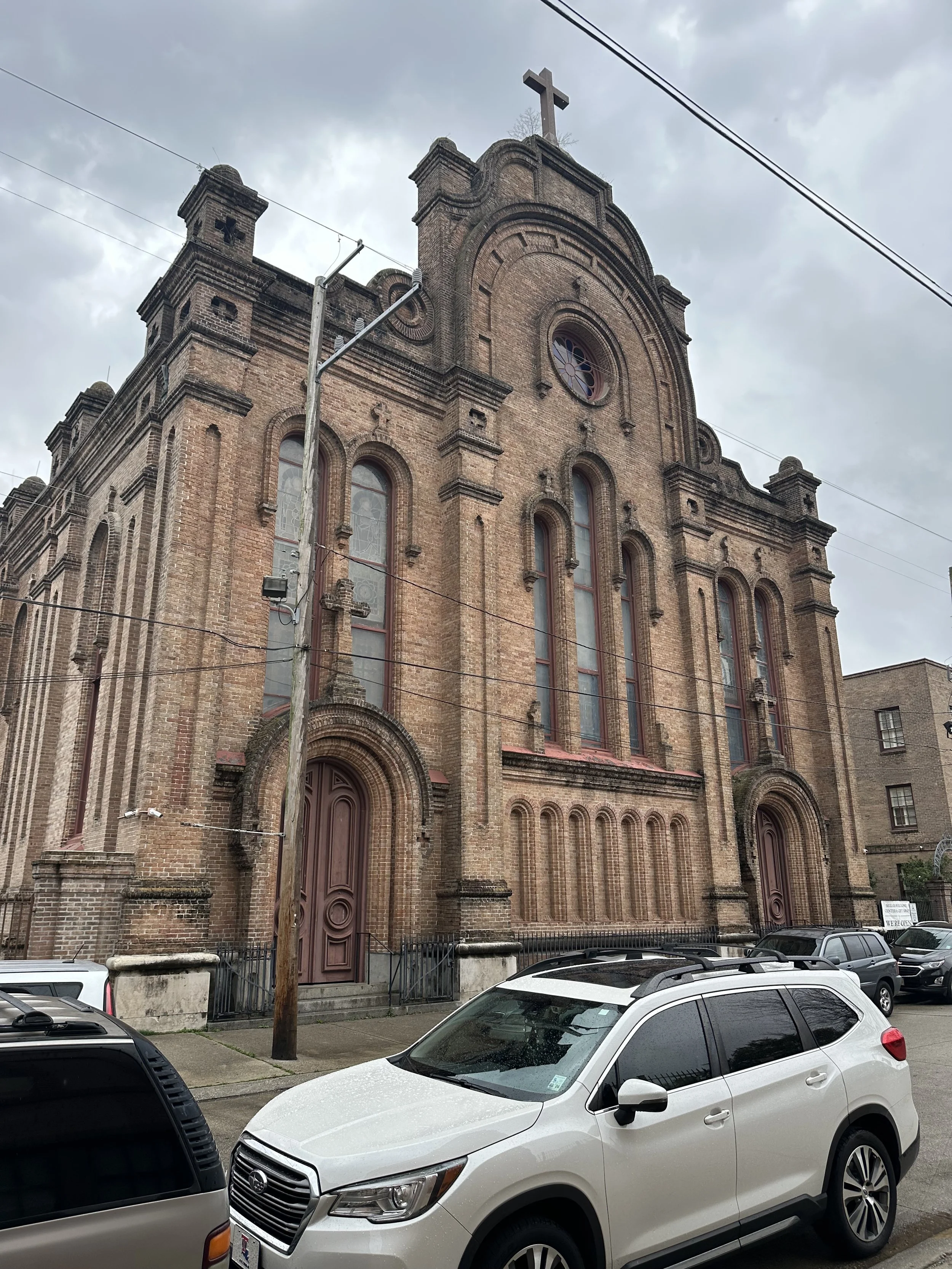 A large historic brick church with tall arched windows, a cross on top, and intricate architectural details, seen on a cloudy day with parked cars in front.