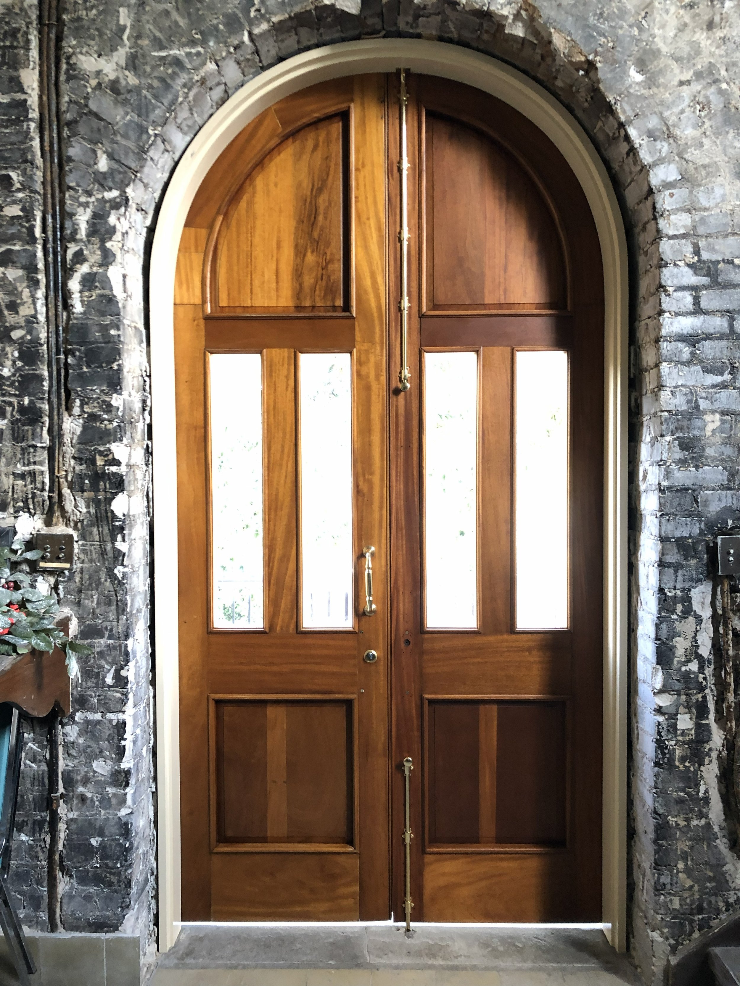Interior of a building under renovation with exposed brick, unfinished walls, and construction materials such as cement bags and wooden planks on the floor, leading towards a doorway with a view of a partially painted green door.