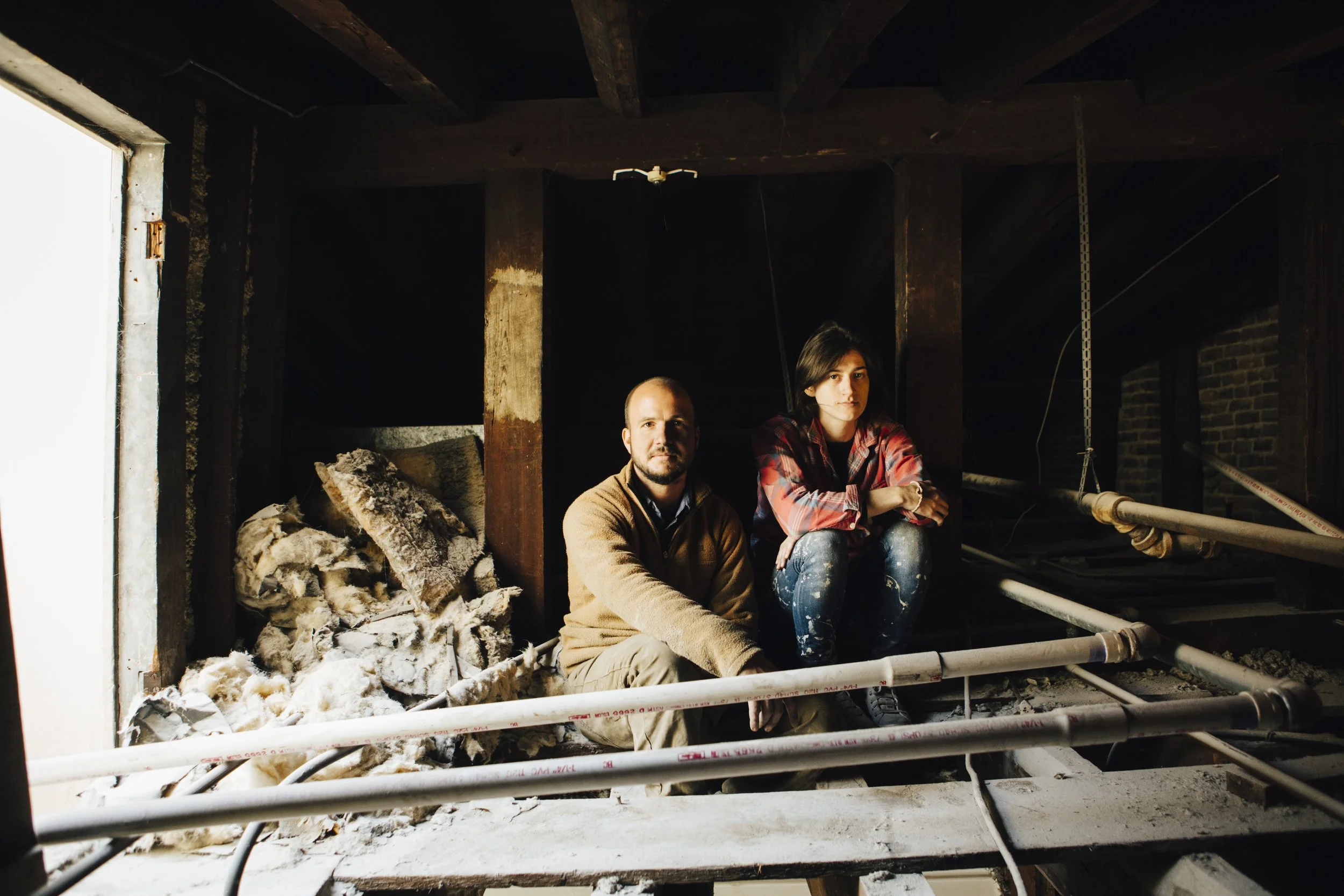Two people sitting on the floor inside a partially constructed or renovated room with exposed pipes, insulation, and wooden beams.