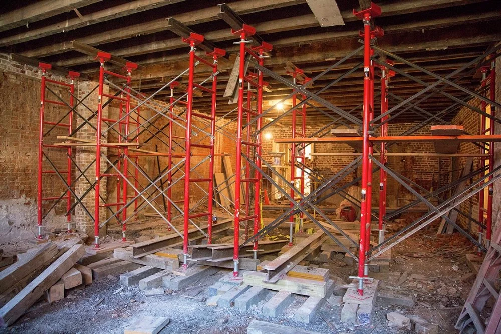 Construction site inside a building with red and black scaffolding structures, wooden planks, and construction debris on the floor.