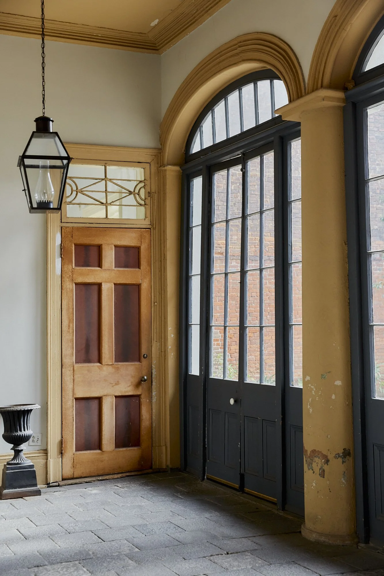 Interior of a historic building with large arched windows, a wooden door, a hanging lantern light, and weathered yellow walls with peeling paint.